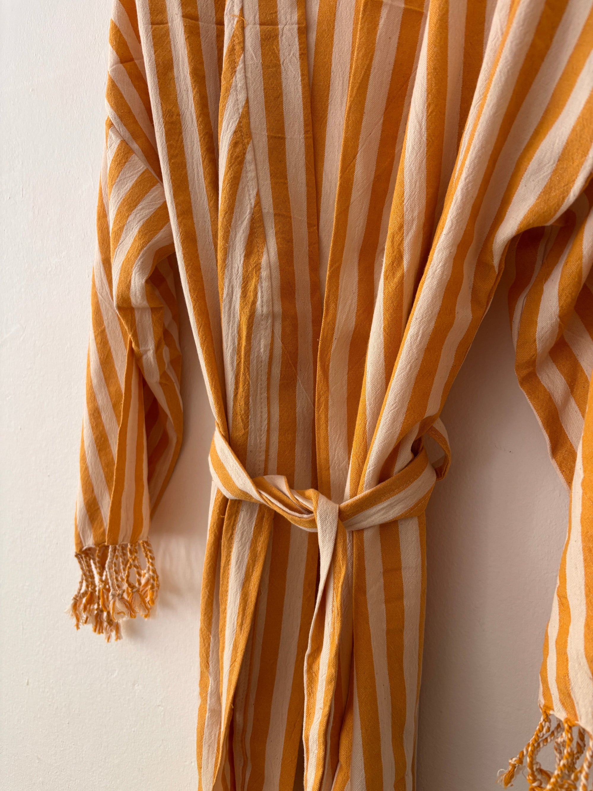 Der KIMONO - MEDITERANE - GELB - WEIß von THE DANISH GIRL INTERIOR mit vertikalen gelb-weißen Streifen, Taillengürtel und Quasten an den Ärmeln hängt an einer weißen Wand.
