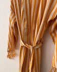 Der KIMONO - MEDITERANE - GELB - WEIß von THE DANISH GIRL INTERIOR mit vertikalen gelb-weißen Streifen, Taillengürtel und Quasten an den Ärmeln hängt an einer weißen Wand.