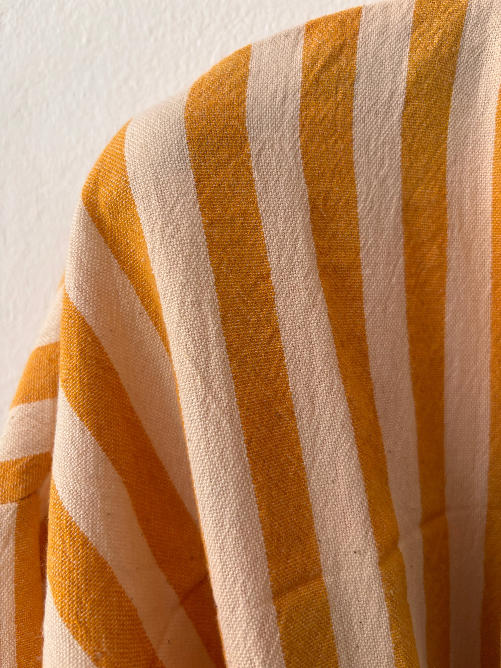 Eine Nahaufnahme des KIMONO - MEDITERANE - GELB - WEIß von THE DANISH GIRL INTERIOR zeigt vertikale gelbe und weiße Streifen, wobei die sanften Falten und die Textur des Stoffes vor einer schlichten hellen Wand sichtbar sind.