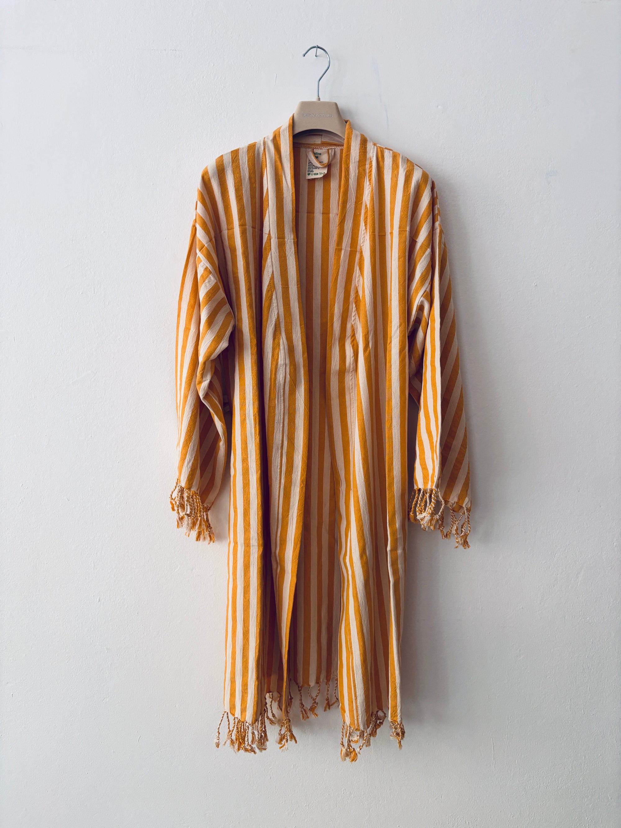 Das KIMONO - MEDITERANE - GELB - WEIß von THE DANISH GIRL INTERIOR hängt auf einem Holzbügel an einer weißen Wand. Lange Ärmel, gelb-weiße Längsstreifen und lose Fransen an den Manschetten und am Saum sorgen für einen entspannten Stil.