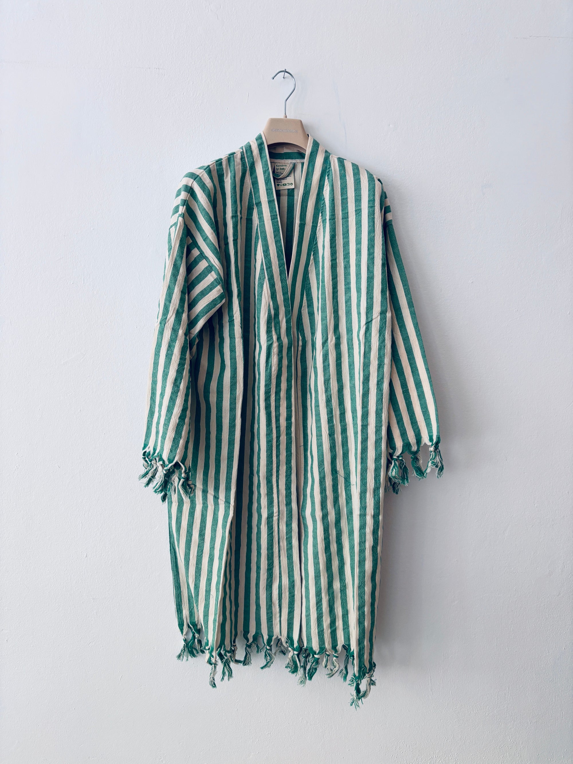 Der KIMONO - MEDITERANE - GRÜN - WEIß von THE DANISH GIRL INTERIOR mit Streifen, langen Ärmeln und Fransen hängt an einem Holzkleiderbügel vor einer schlichten weißen Wand.