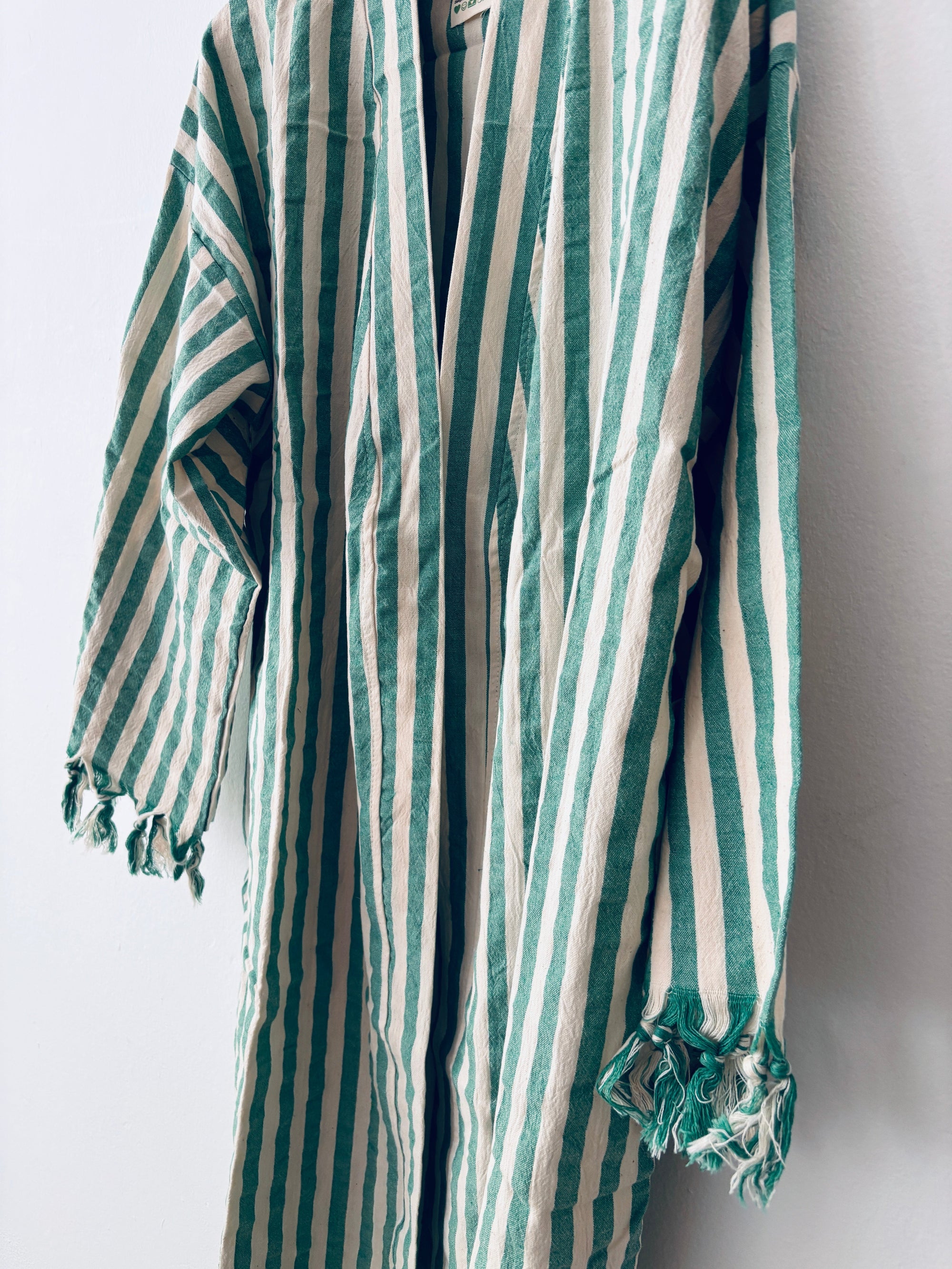 Der KIMONO - MEDITERANE - GRÜN - WEIß von THE DANISH GIRL INTERIOR mit langen Ärmeln und Fransen an den Bündchen besticht durch grüne und weiße Streifen für einen frischen, stilvollen Look vor weißer Wand.