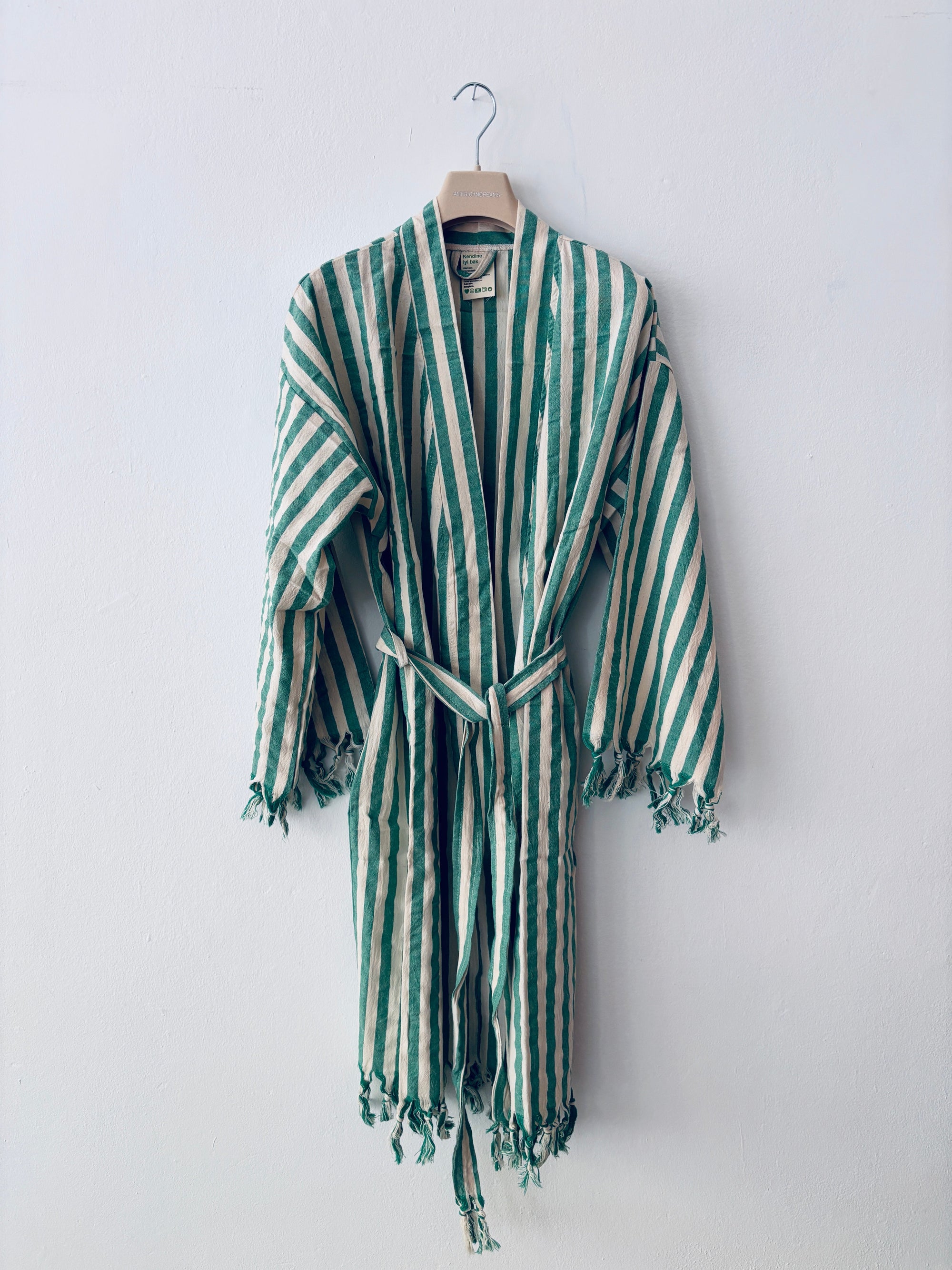 Der KIMONO - MEDITERANE - GRÜN - WEIß von THE DANISH GIRL INTERIOR ist ein grün-weiß gestreifter Bademantel mit langen Ärmeln, Taillengürtel und geflochtenen Rändern, der auf einem Holzbügel vor einer schlichten weißen Wand hängt.