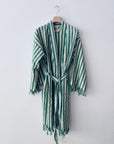 Der KIMONO - MEDITERANE - GRÜN - WEIß von THE DANISH GIRL INTERIOR ist ein grün-weiß gestreifter Bademantel mit langen Ärmeln, Taillengürtel und geflochtenen Rändern, der auf einem Holzbügel vor einer schlichten weißen Wand hängt.