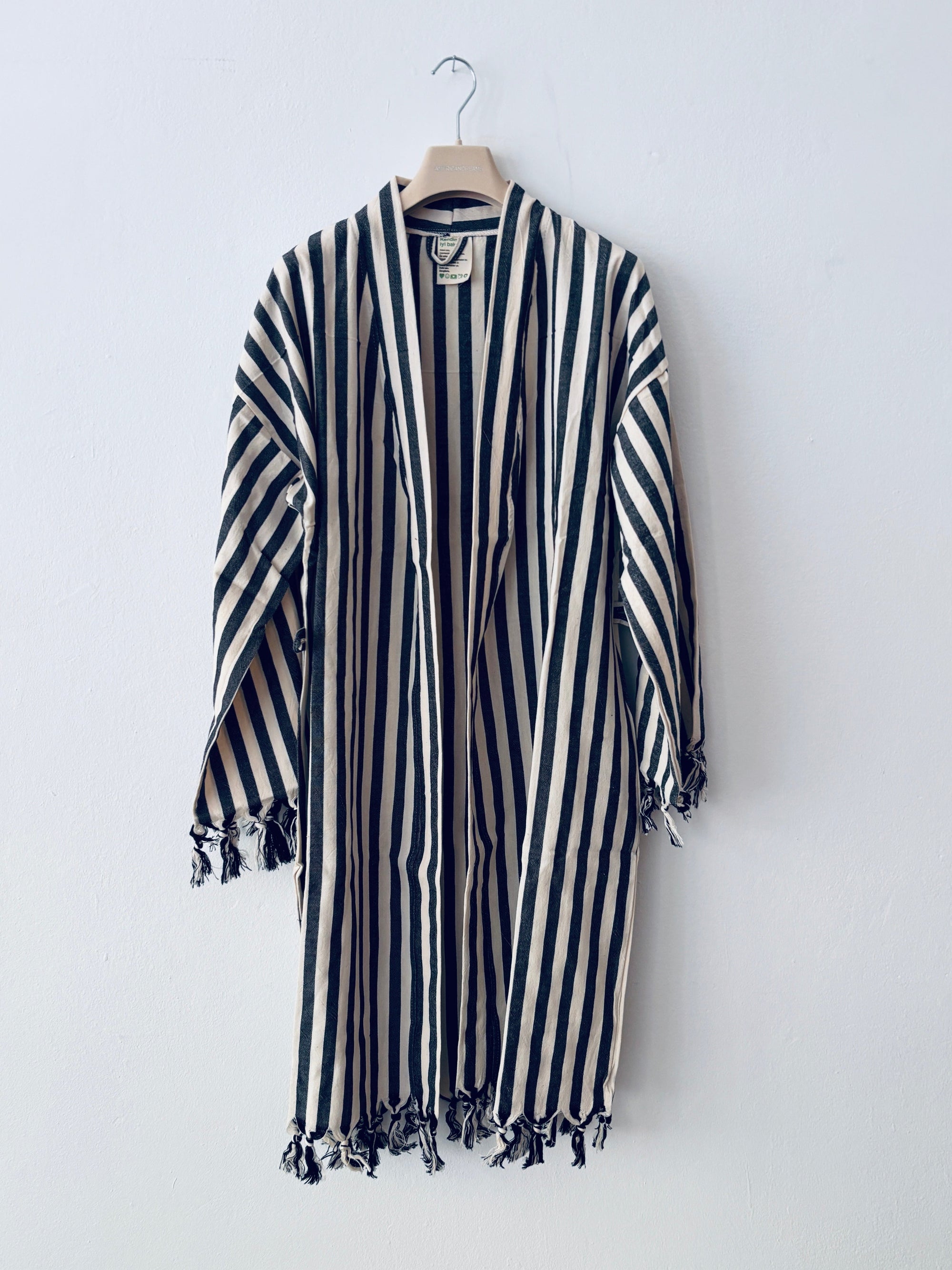 Der KIMONO - MEDITERANE - SCHWARZ - WEIß von THE DANISH GIRL INTERIOR ist ein langer, locker sitzender Kimono mit schwarzen und weißen Längsstreifen und Fransen an den Ärmeln und am Saum, der auf einem Holzbügel vor einer weißen Wand gezeigt wird.