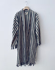 Der KIMONO - MEDITERANE - SCHWARZ - WEIß von THE DANISH GIRL INTERIOR ist ein langer, locker sitzender Kimono mit schwarzen und weißen Längsstreifen und Fransen an den Ärmeln und am Saum, der auf einem Holzbügel vor einer weißen Wand gezeigt wird.