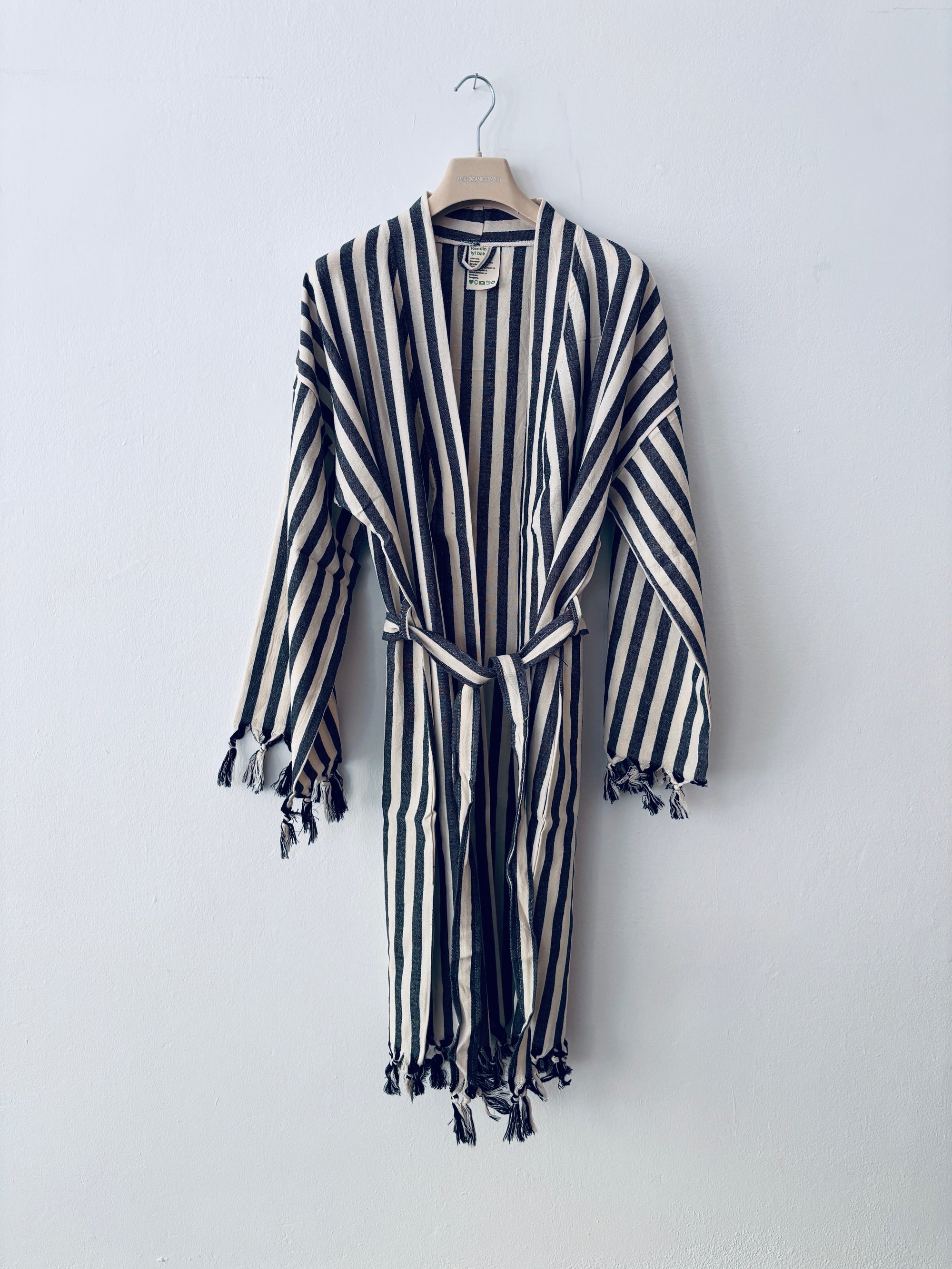 Der KIMONO - MEDITERANE - SCHWARZ - WEIß von THE DANISH GIRL INTERIOR hängt gestreift und mit Quasten an Ärmeln und Saum an einem Holzkleiderbügel vor einer weißen Wand.