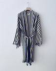 Der KIMONO - MEDITERANE - SCHWARZ - WEIß von THE DANISH GIRL INTERIOR hängt gestreift und mit Quasten an Ärmeln und Saum an einem Holzkleiderbügel vor einer weißen Wand.