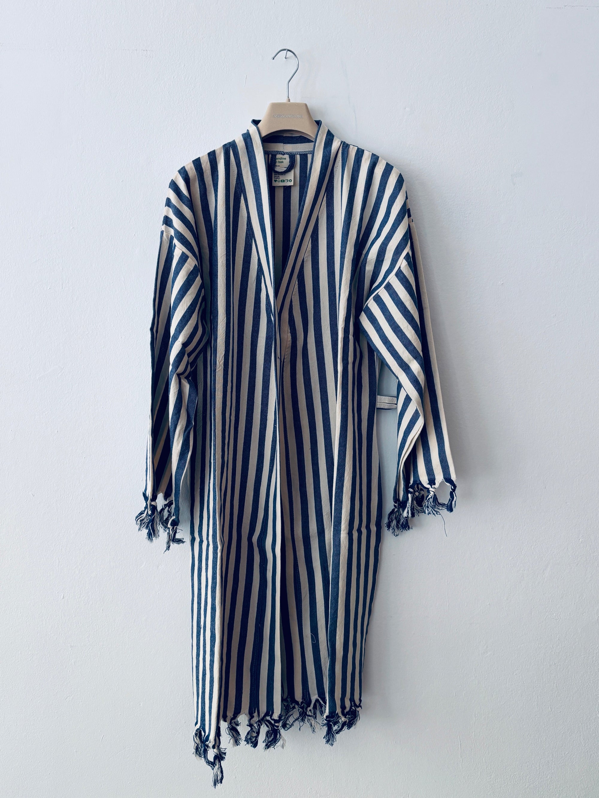 Das Kleidungsstück KIMONO - MEDITERANE - BLAU - WEIß von THE DANISH GIRL INTERIOR mit blauen und weißen Längsstreifen, langen Ärmeln und Quasten an den Rändern wird auf einem Holzbügel vor einer schlichten weißen Wand präsentiert.
