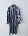 Das Kleidungsstück KIMONO - MEDITERANE - BLAU - WEIß von THE DANISH GIRL INTERIOR mit blauen und weißen Längsstreifen, langen Ärmeln und Quasten an den Rändern wird auf einem Holzbügel vor einer schlichten weißen Wand präsentiert.