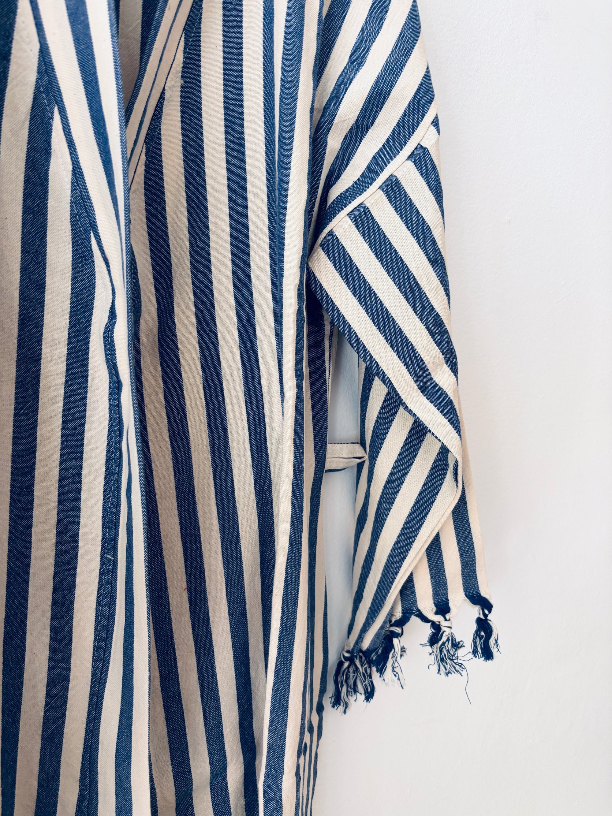 Eine Nahaufnahme des KIMONO - MEDITERANE - BLAU - WEIß von THE DANISH GIRL INTERIOR mit blauen und weißen vertikalen Streifen und einem ausgefransten Ärmel, der vor einem weißen Hintergrund hängt.