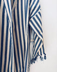 Eine Nahaufnahme des KIMONO - MEDITERANE - BLAU - WEIß von THE DANISH GIRL INTERIOR mit blauen und weißen vertikalen Streifen und einem ausgefransten Ärmel, der vor einem weißen Hintergrund hängt.
