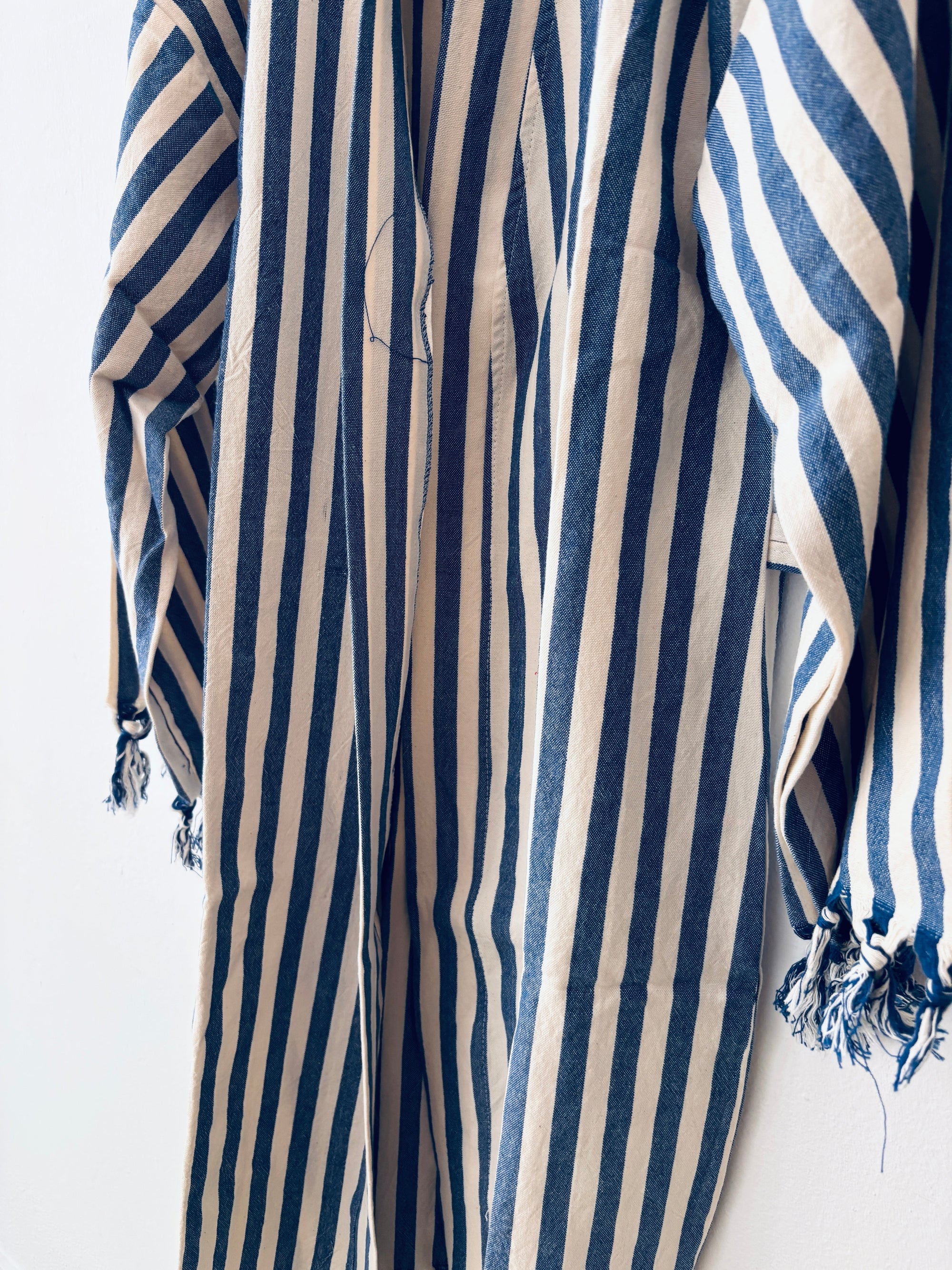 Eine Nahaufnahme des KIMONO - MEDITERANE - BLAU - WEIß von THE DANISH GIRL INTERIOR zeigt die blau-weißen Längsstreifen, lange Ärmel mit ausgefransten Manschetten und einen locker hängenden, leichten Baumwollmantelstoff für einen entspannten Look.