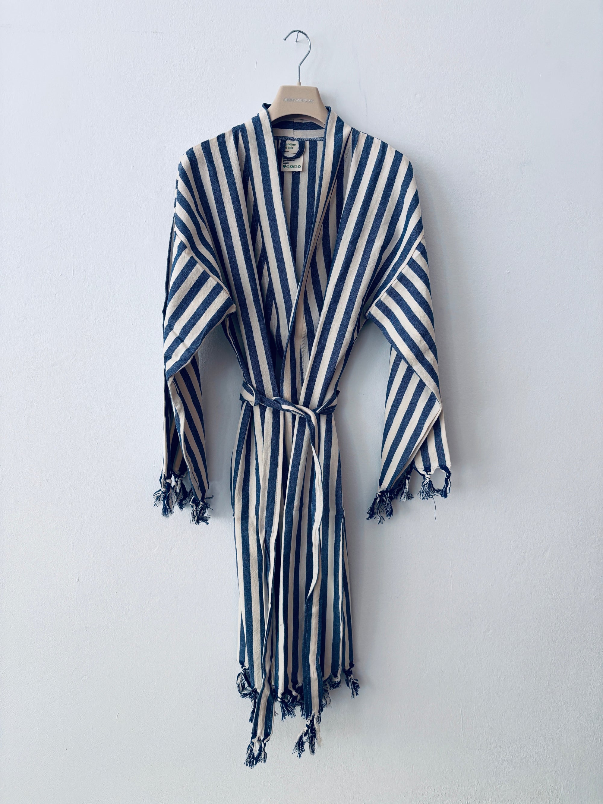 Der KIMONO - MEDITERANE - BLAU - WEIß von THE DANISH GIRL INTERIOR aus Baumwolle mit blauen und weißen Streifen, Gürtel sowie Fransen an Ärmeln und Saum hängt an einem Holzbügel vor einer weißen Wand.