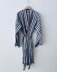 Der KIMONO - MEDITERANE - BLAU - WEIß von THE DANISH GIRL INTERIOR aus Baumwolle mit blauen und weißen Streifen, Gürtel sowie Fransen an Ärmeln und Saum hängt an einem Holzbügel vor einer weißen Wand.