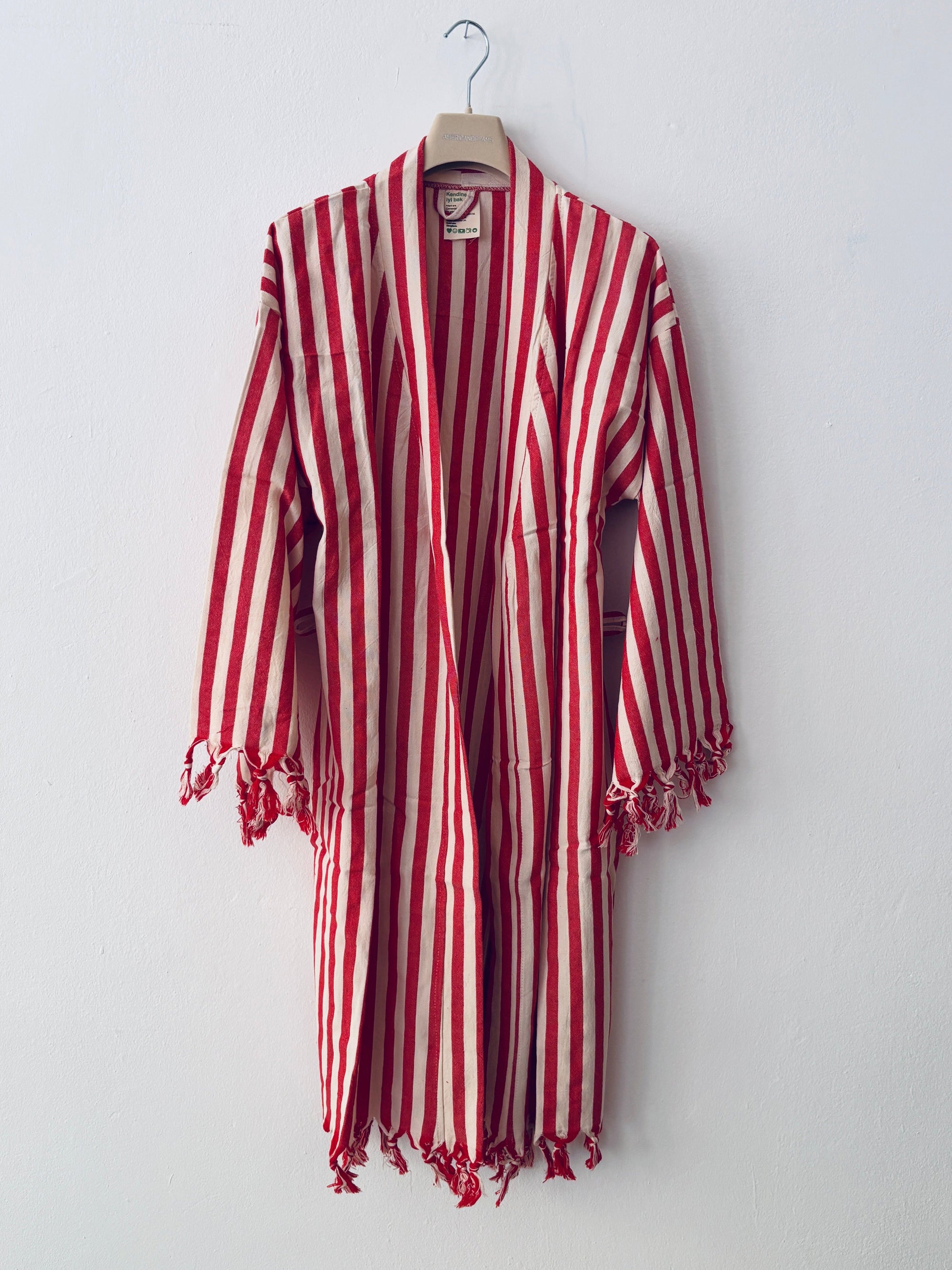 Der KIMONO - MEDITERANE - ROT - WEIß von THE DANISH GIRL INTERIOR mit roten und weißen Streifen, langen Ärmeln und Fransen hängt an einem Haken vor einer schlichten weißen Wand.