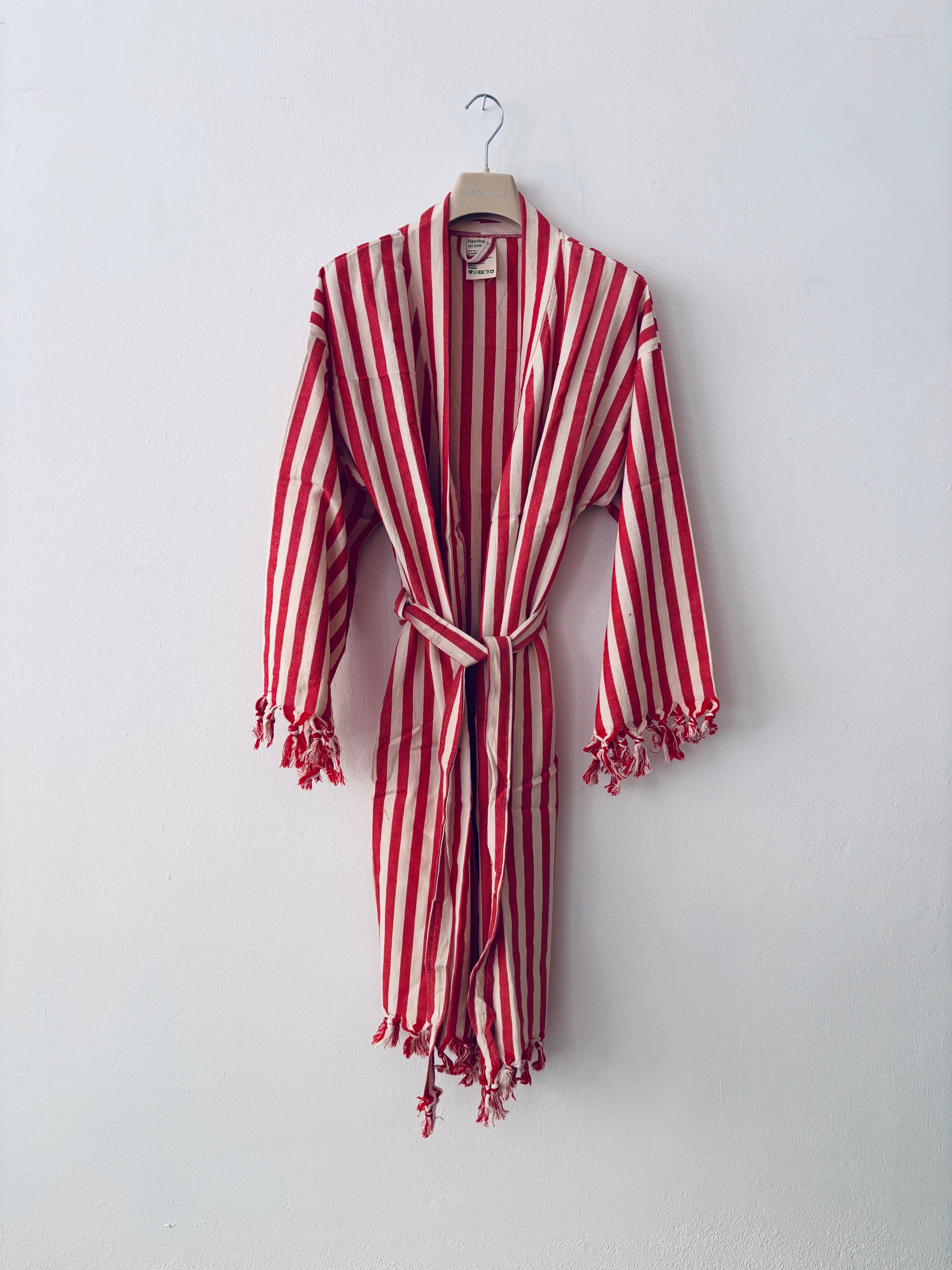 Der KIMONO - MEDITERANE - ROT - WEIß von THE DANISH GIRL INTERIOR mit Gürtel und Quasten hängt an einem Holzkleiderbügel vor einer weißen Wand.