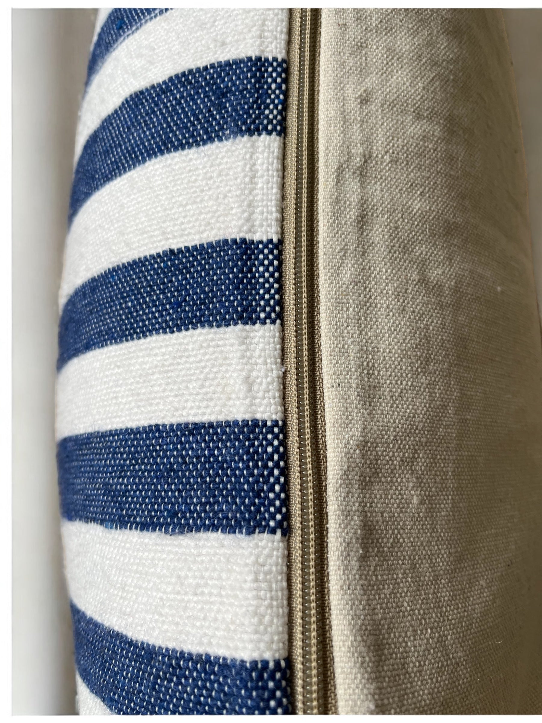 Nahaufnahme des LEY! Kissen - Hellas 60x60. Man sieht blaue und weiße Streifen auf der einen Seite, einfarbiges Beige auf der anderen Seite und einen beigen Reißverschluss, der vertikal zwischen den beiden Stoffen verläuft.