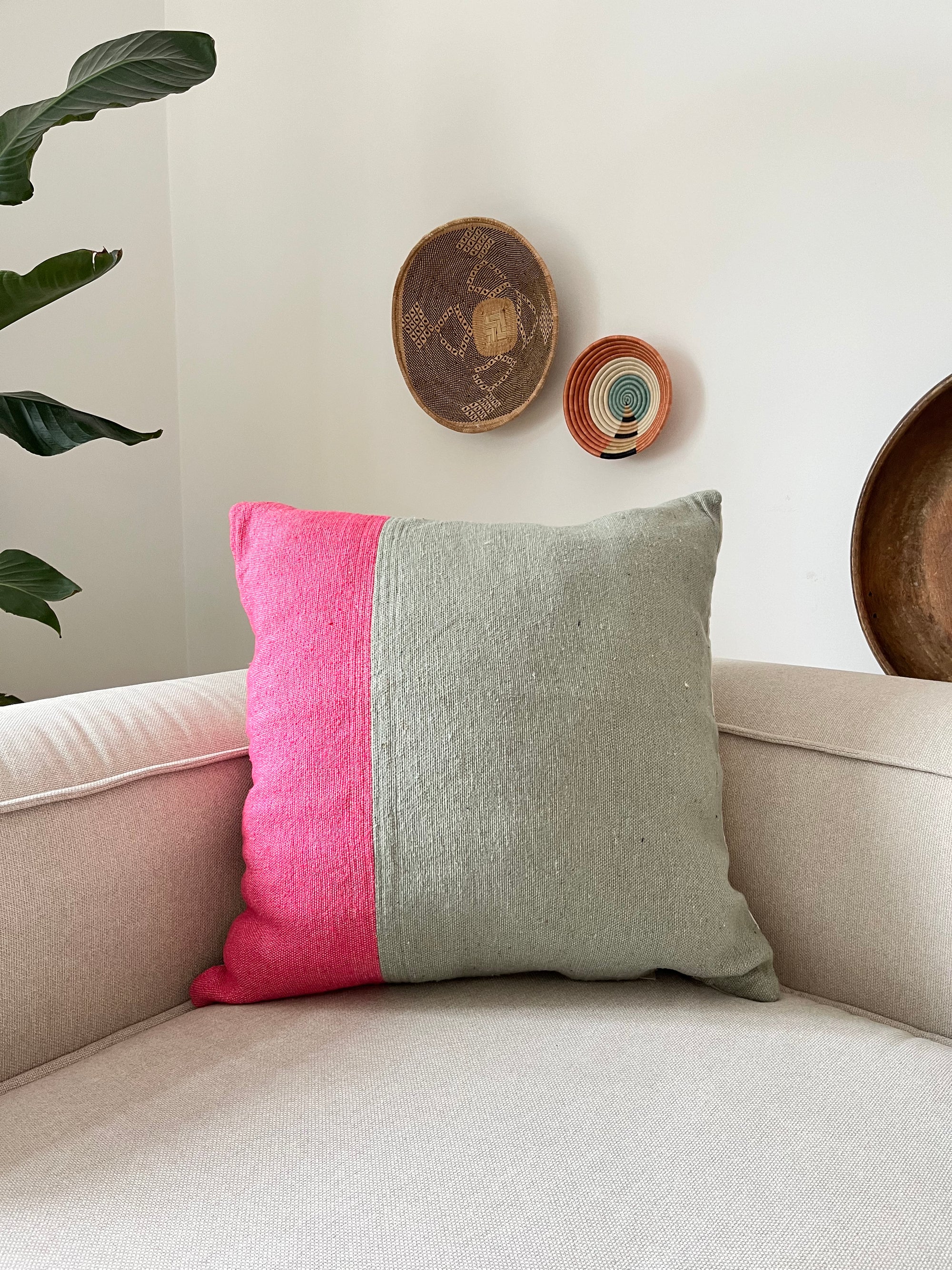 Auf einem beigen Sessel liegt das LEY! Kissen - Merva 60x60, halb grau, halb hellrosa. Drei gewebte Wandbehänge und eine große Pflanze bringen Stil in die weiße Wand dahinter.
