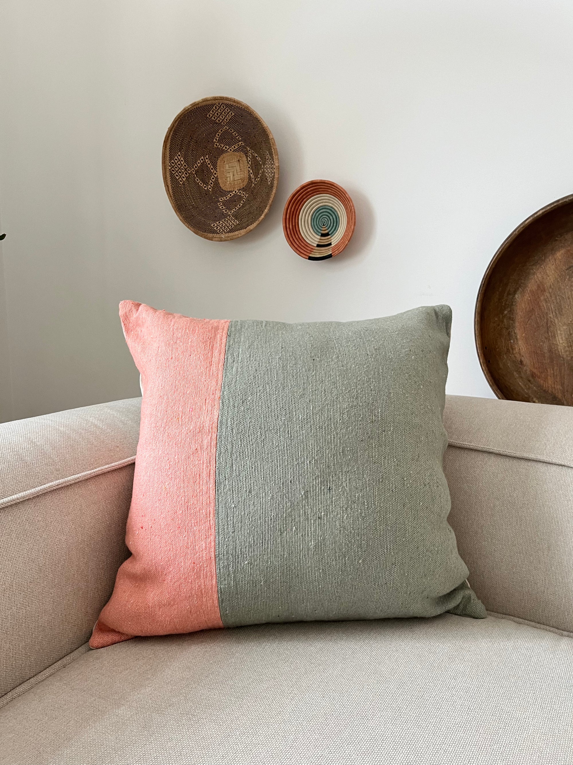 EIN LEY! Kissen - Maila 60x60 in rosa und hellgrauem Farbblock ruht auf einem beigen Stuhl, an der weißen Wand dahinter hängen drei handgefertigte Marrakesch-Flechtkörbe.