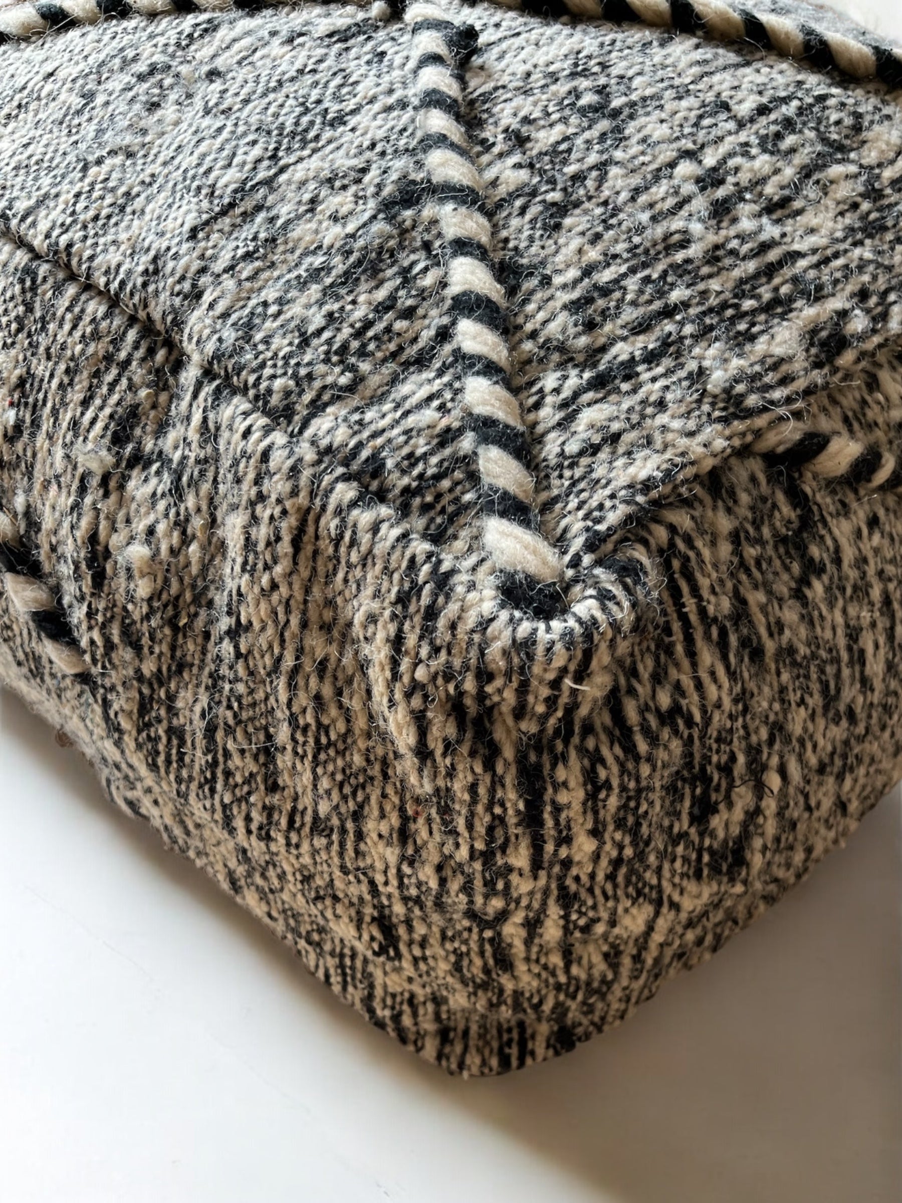 Eine Nahaufnahme des Kleinen Bodenkissens - Mica Mini von LEY!, ein schwarz-weißer Hocker mit diagonalen Seilakzenten, handgefertigt aus hochwertiger Schurwolle, der auf einem glatten, hellen Boden liegt.