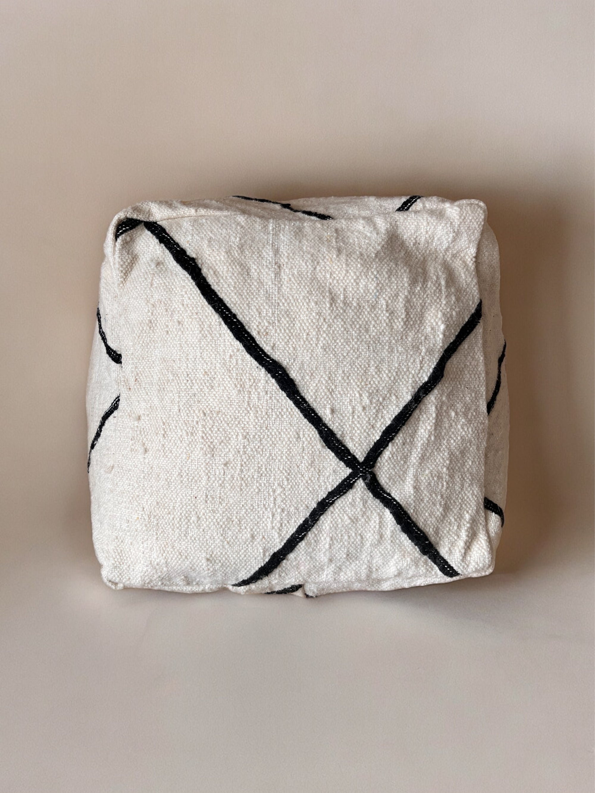 Das "Kleine Bodenkissen - Meli Mini" von LEY! ist ein quadratisches, off-white handgefertigtes Kissen aus Schurwolle mit schwarzen, diagonal verlaufenden Linien im X-Muster - präsentiert vor neutralem Hintergrund.