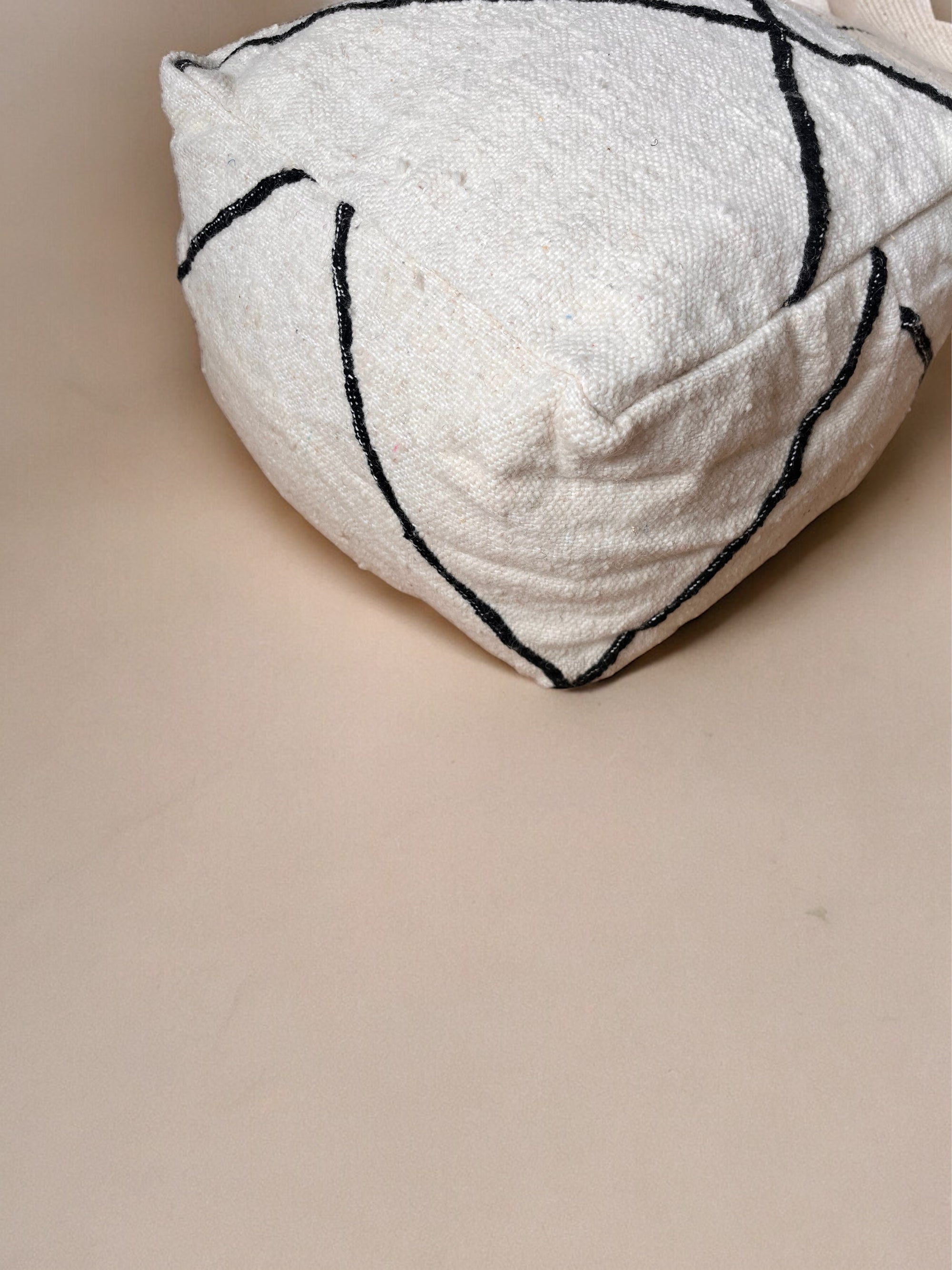 Eine Nahaufnahme des LEY! Kleines Bodenkissen - Meli Mini, ein handgefertigtes, weißes, strukturiertes Kissen mit schwarzen, geschwungenen Linien, das auf einer glatten, beigen Oberfläche ruht.