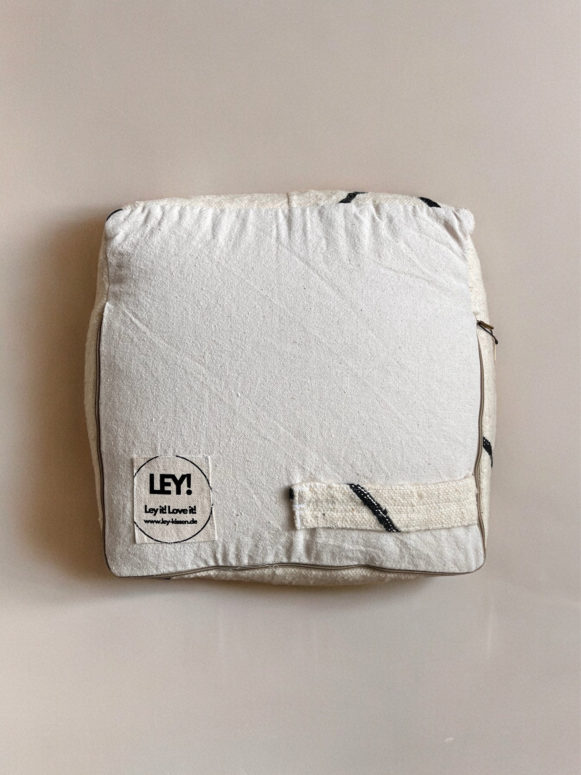 Ein weißes, stoffbespanntes Quadrat "Kleines Bodenkissen - Meli Mini" von LEY!, handgefertigt aus Schurwolle, hat einen kleinen Aufnäher mit "LEY! Lay it! Love it!" in einer Ecke und ist auf einem neutralen Hintergrund abgebildet.
