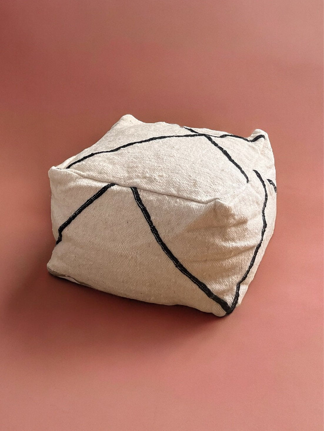 Ein beigefarbener, strukturierter, quadratischer Hocker mit schwarzen, diagonalen Linien liegt auf einer gedämpften rosa Oberfläche.