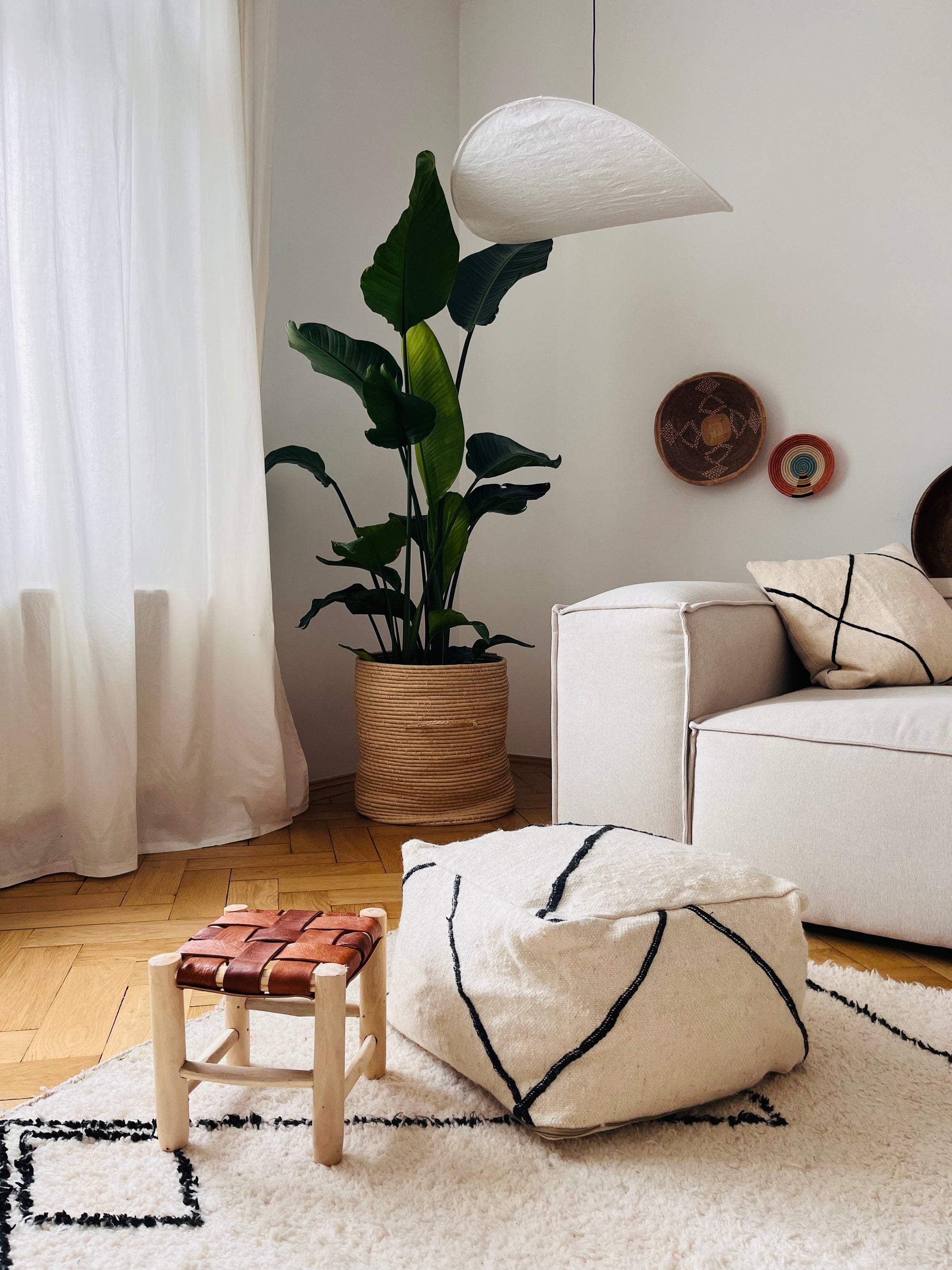 Ein gemütliches, modernes Wohnzimmer mit einem beigen Sofa, geometrischen Kissen, einer großen Topfpflanze und dem LEY! Kleines Bodenkissen - Meli Mini. Das Licht fällt durch die transparenten Vorhänge und hebt die natürlichen Farbtöne und Texturen des Raums hervor.