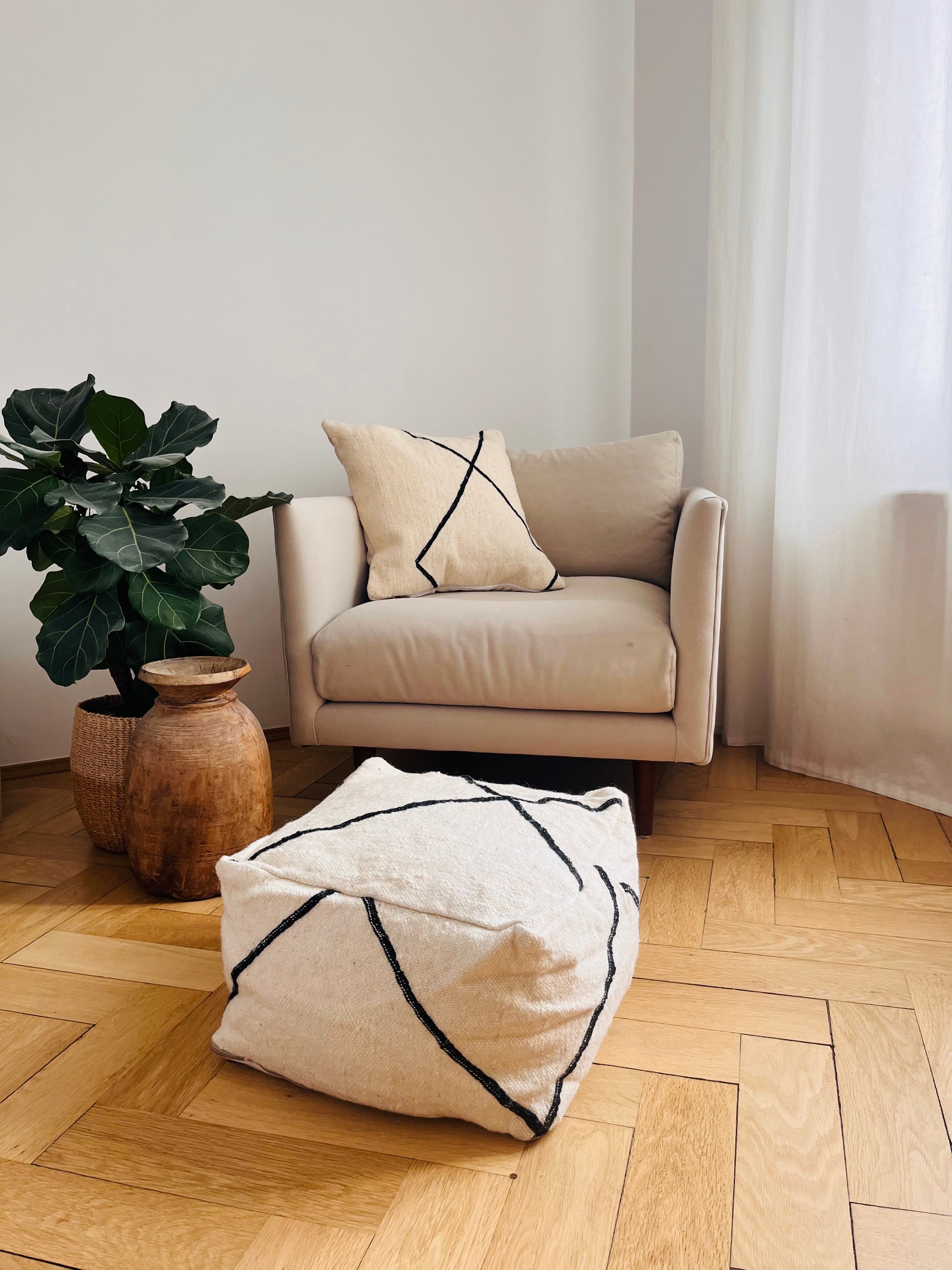 Ein gemütlicher beiger Sessel mit einem geometrischen Kissen steht auf einem Holzboden neben einer blattreichen Topfpflanze, einer rustikalen Vase und dem LEY! Kleines Bodenkissen - Meli Mini in Schurwolle in einem hellen, minimalistischen Raum mit weißen Wänden und Vorhängen.