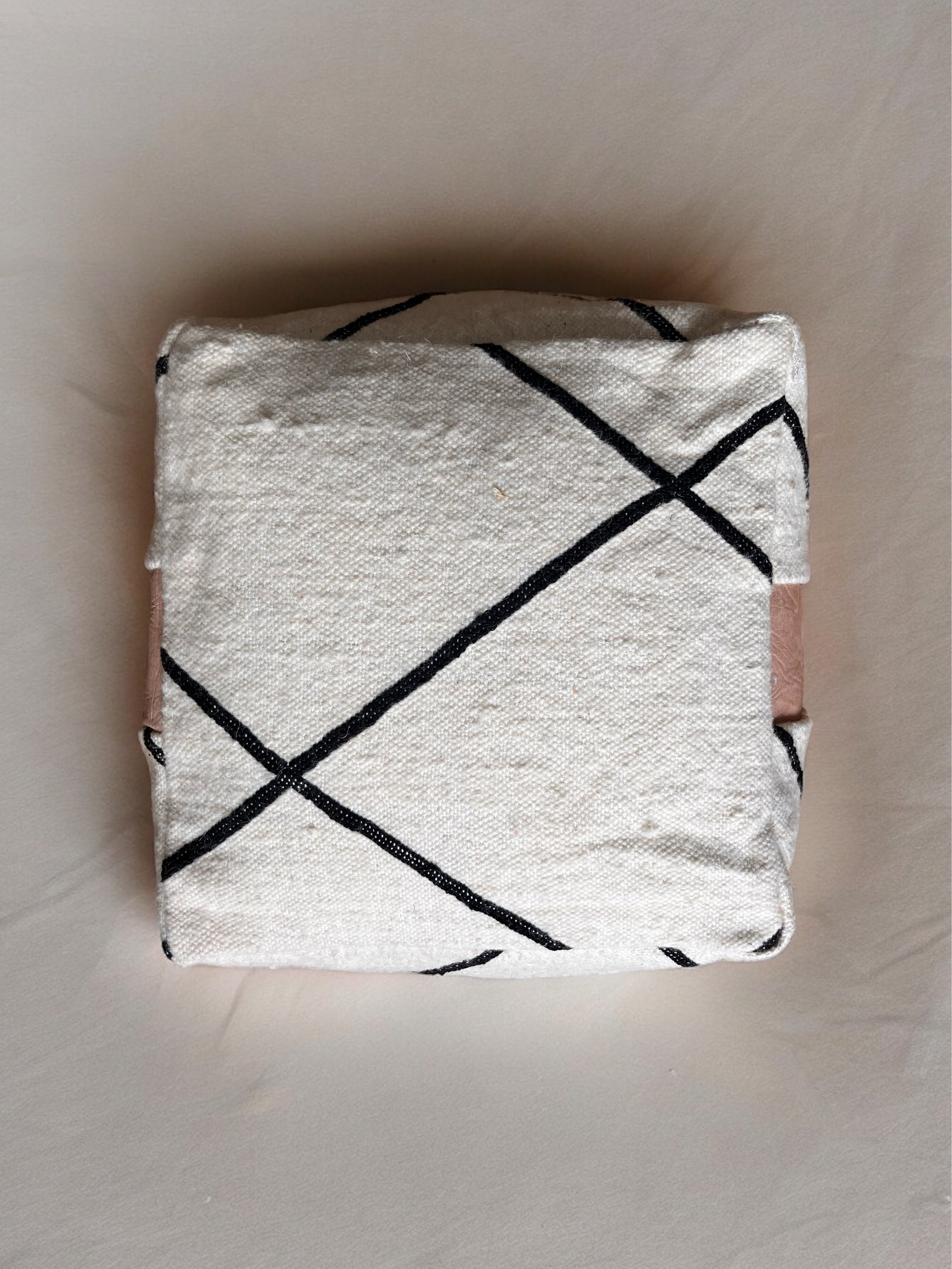 Das Kleine Bodenkissen - Milu Mini von LEY! ist ein quadratisches Kissen mit weißem Stoffbezug und schlichten schwarzen Kreuzlinien, das in Handarbeit gefertigt wurde und auf einer neutralen, hellen Oberfläche steht.