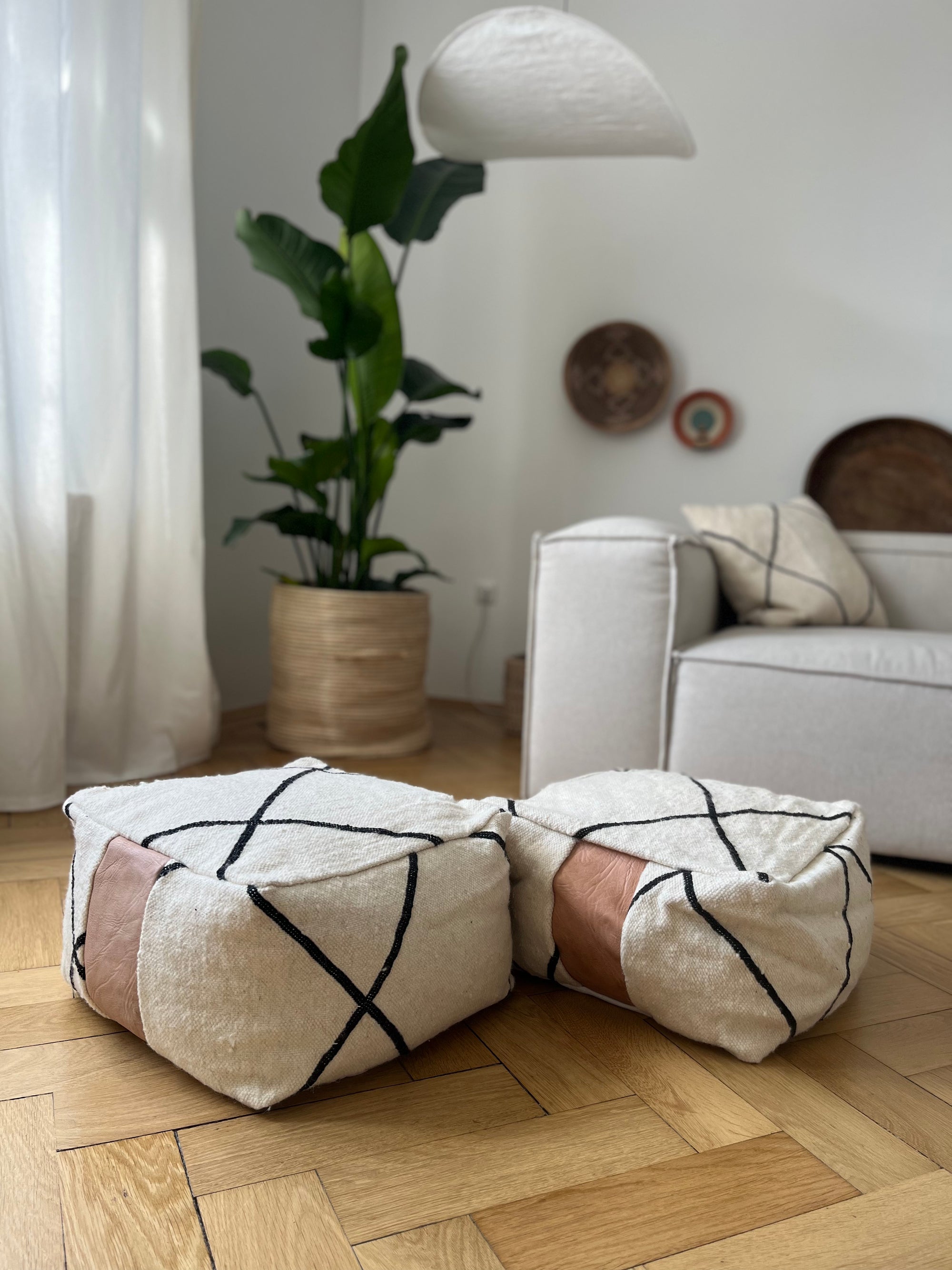 Zwei beigefarbene Poufs mit schwarzen geometrischen Linien und braunen Akzenten stehen in einem hellen, modernen Wohnzimmer auf einem Holzboden. Eine große Topfpflanze, ein weißes Sofa, Wandkunst und sanftes natürliches Licht vervollständigen das gemütliche Ambiente.