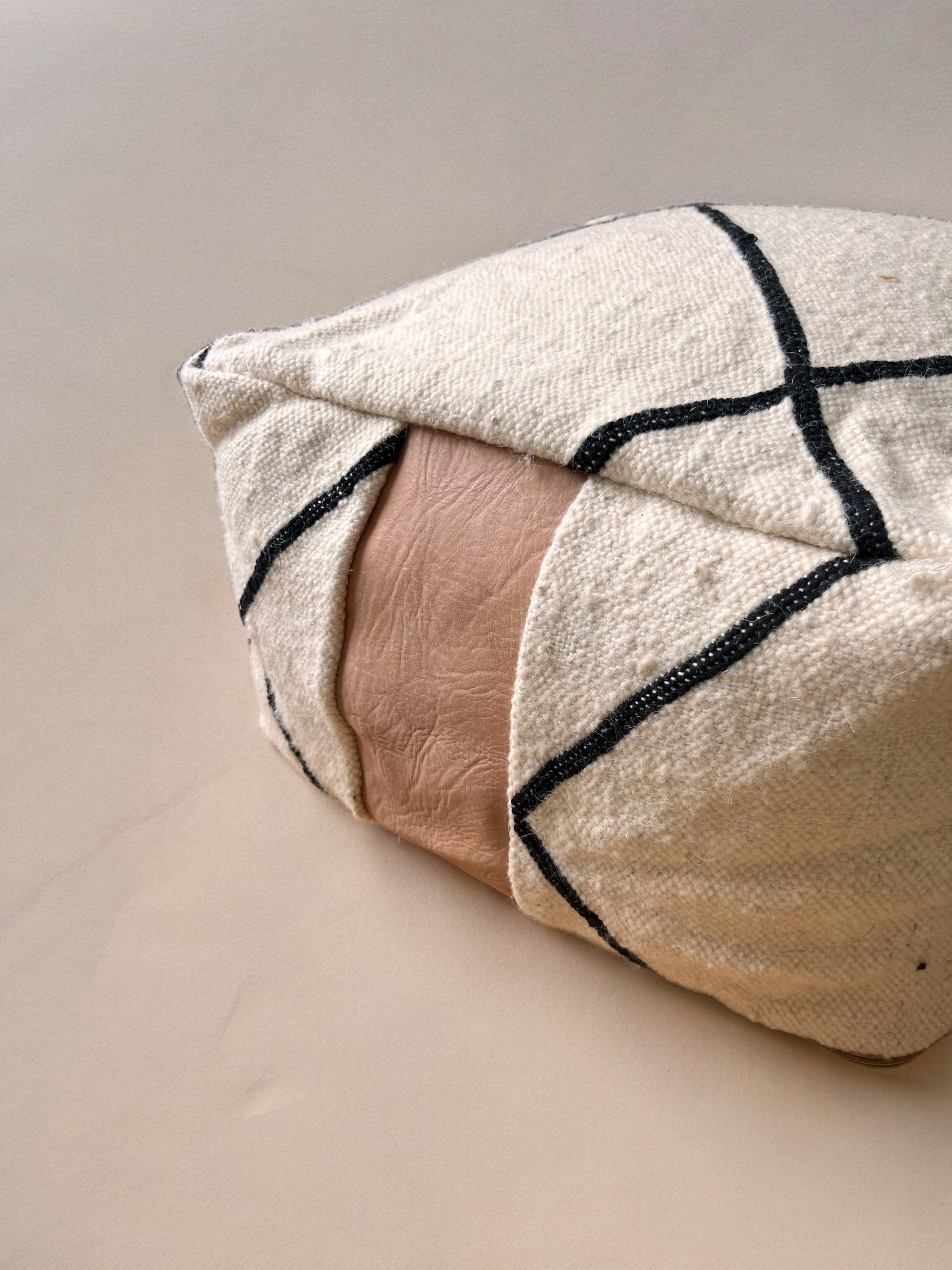Eine Nahaufnahme des LEY! Kleines Bodenkissen - Milu Mini zeigt cremefarbene reine Wolle mit schwarzen geometrischen Linien und einer zentralen hellbraunen Lederplatte, handgefertigt und vor einem neutralen Hintergrund.