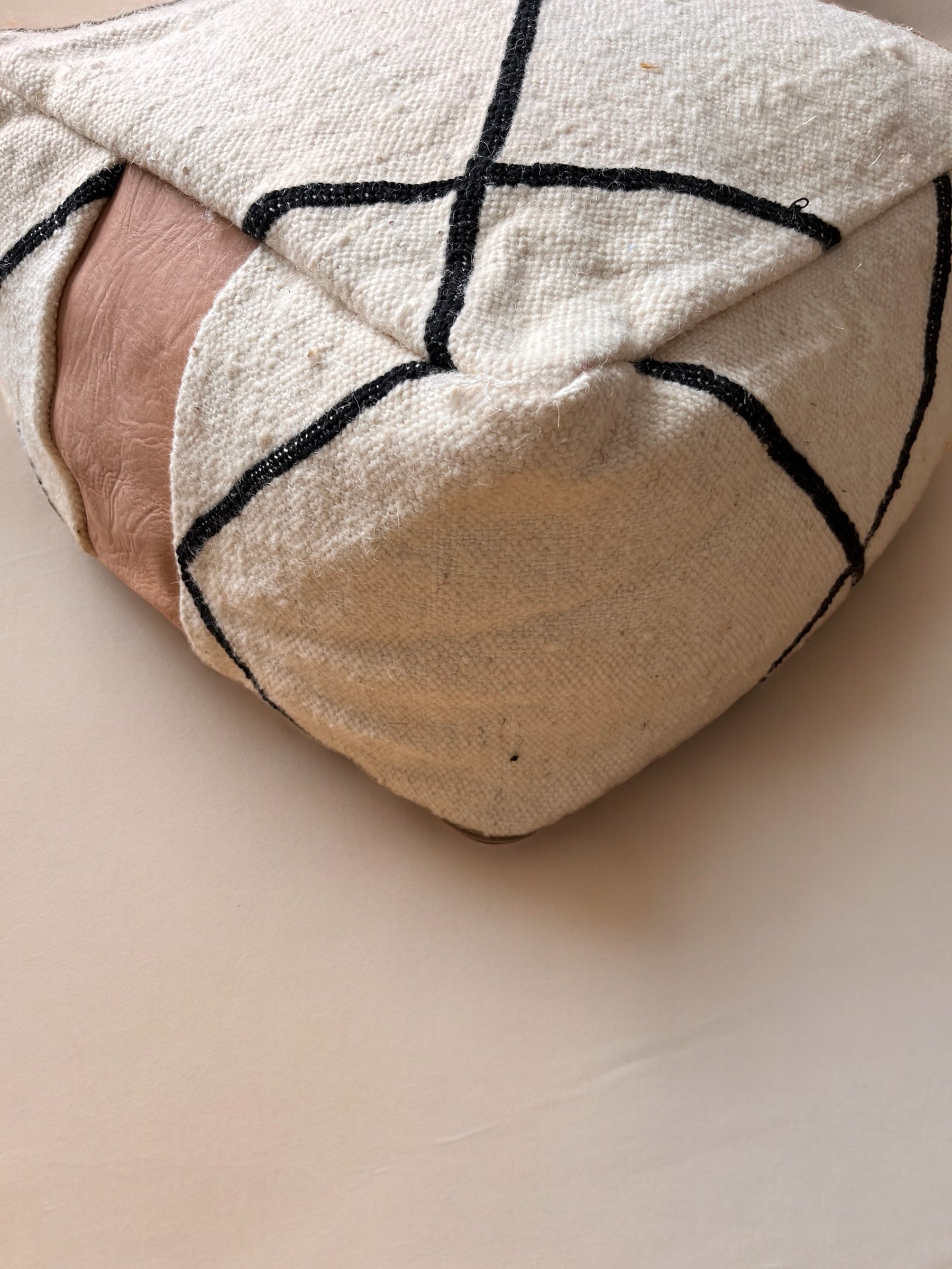 Eine Nahaufnahme des LEY! Kleines Bodenkissen - Milu Mini zeigt seinen weichen, handgefertigten Stoff, das schwarze, geometrische Muster und den braunen Lederakzent, der auf einer hellbeigen Oberfläche ruht.