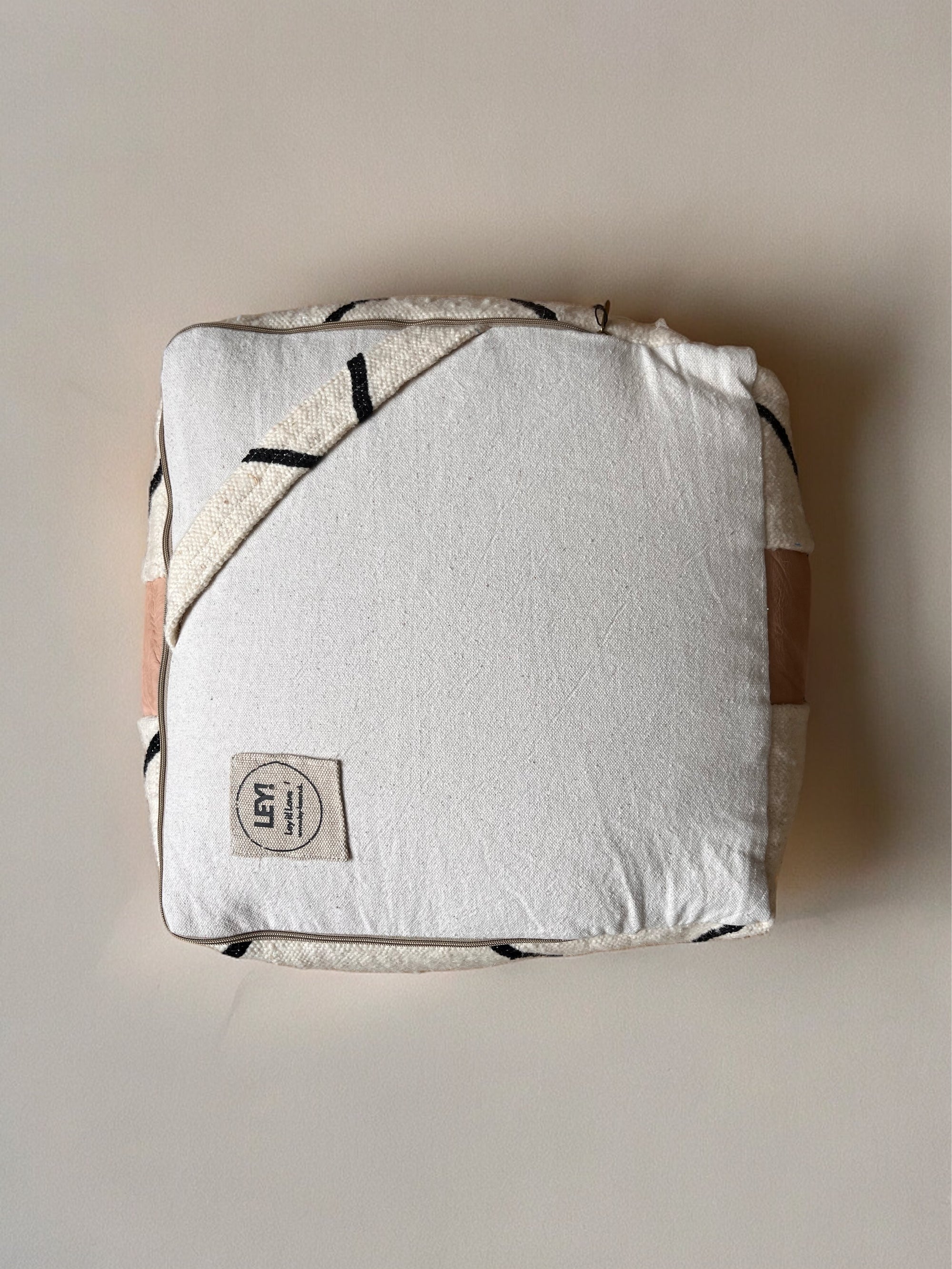 Ein sauber gefaltetes cremefarbenes Kleines Bodenkissen - Milu Mini von LEY!, reine Wolle, mit gesticktem geometrischem Muster, Stoffband und kleinem "LEY!"-Etikett, platziert auf einer glatten hellen Oberfläche.