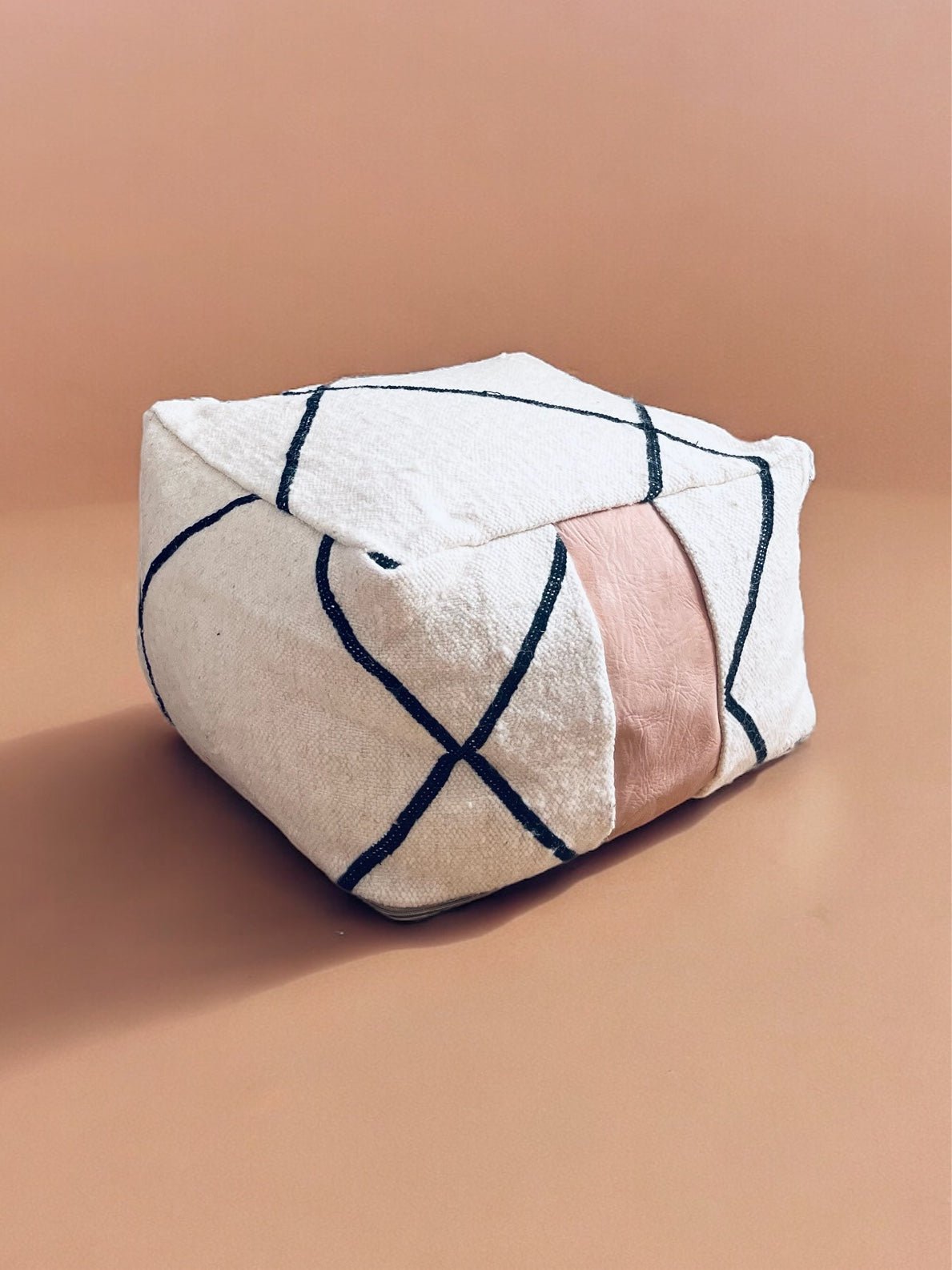 Das kleine Bodenkissen "Milu Mini" von LEY! aus reiner Wolle ist cremefarben, quadratisch, zeigt schwarze Diagonallinien und einen breiten, blassrosa Mittelstreifen - präsentiert auf heller Oberfläche vor passendem Hintergrund.