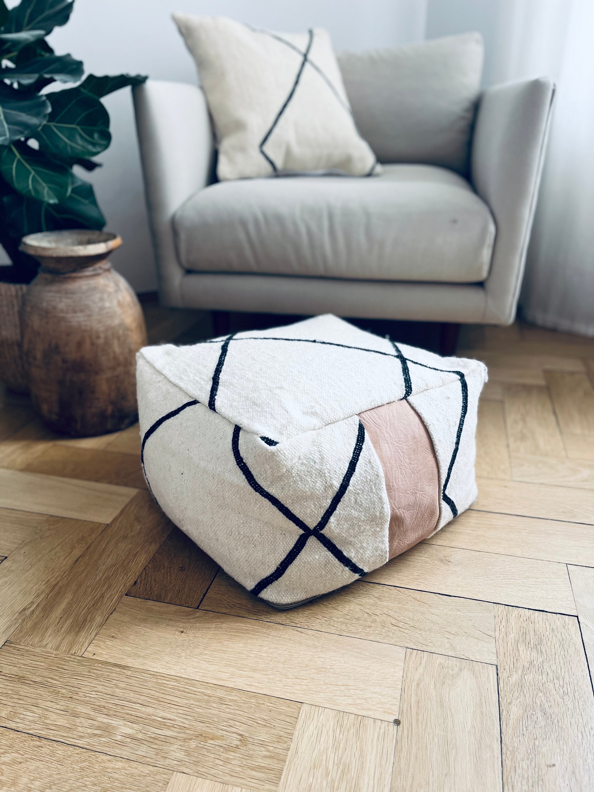 Ein modernes Wohnzimmer mit einem weißen Sessel und Kissen, einer Holzvase und dem LEY! Kleines Bodenkissen - Milu Mini, handgefertigt aus reiner Wolle mit schwarzen Linien und einem beigen Streifen, ruht auf einem hellen Holzboden mit Fischgrätmuster.