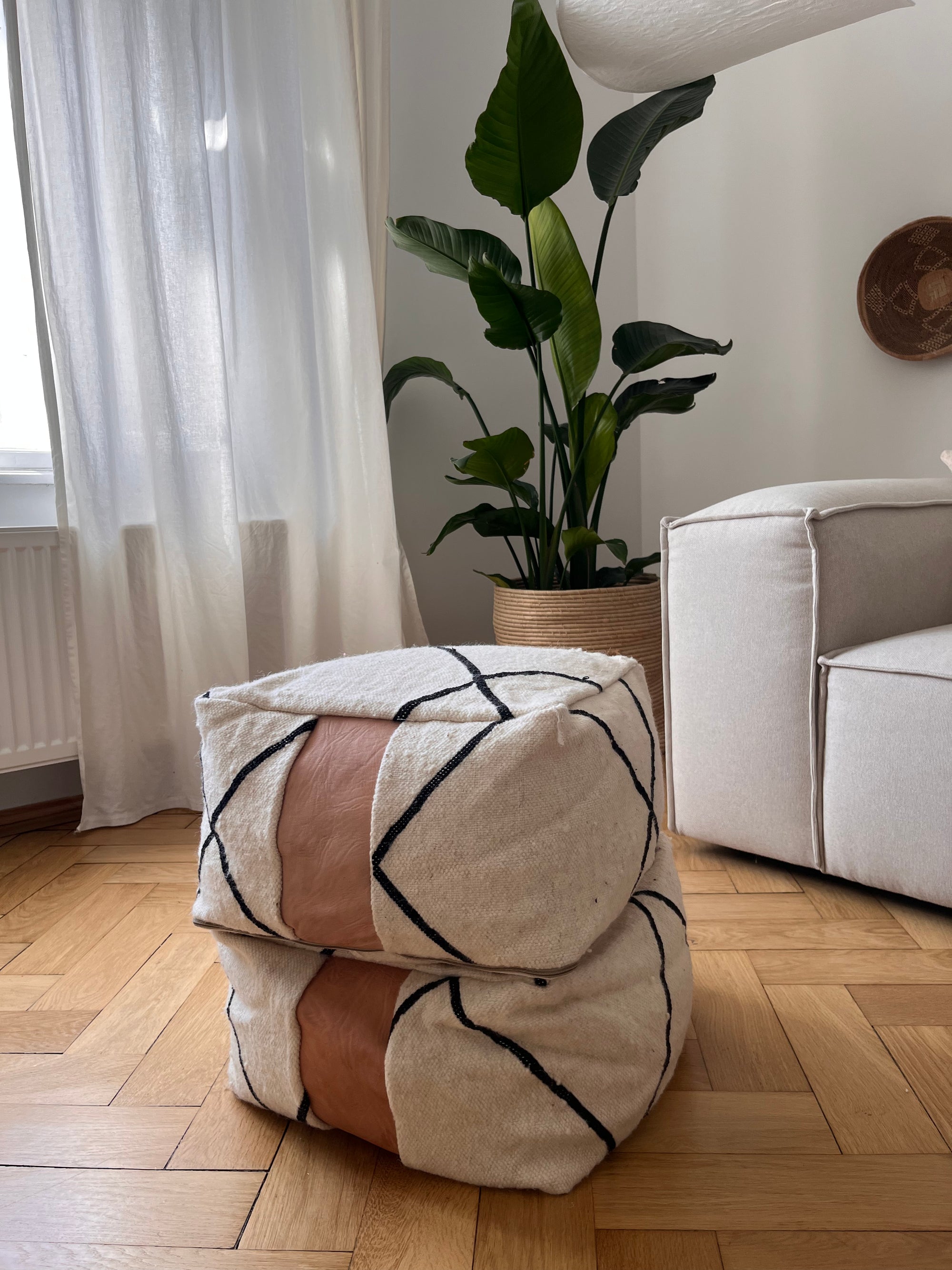 Zwei beige "Kleines Bodenkissen - Milu Mini" von LEY! aus reiner Wolle mit schwarzen Linien und braunen Lederakzenten sind auf Holzboden neben einer grünen Pflanze, weißen Vorhängen und einem Sofa im modernen Wohnzimmer gestapelt.