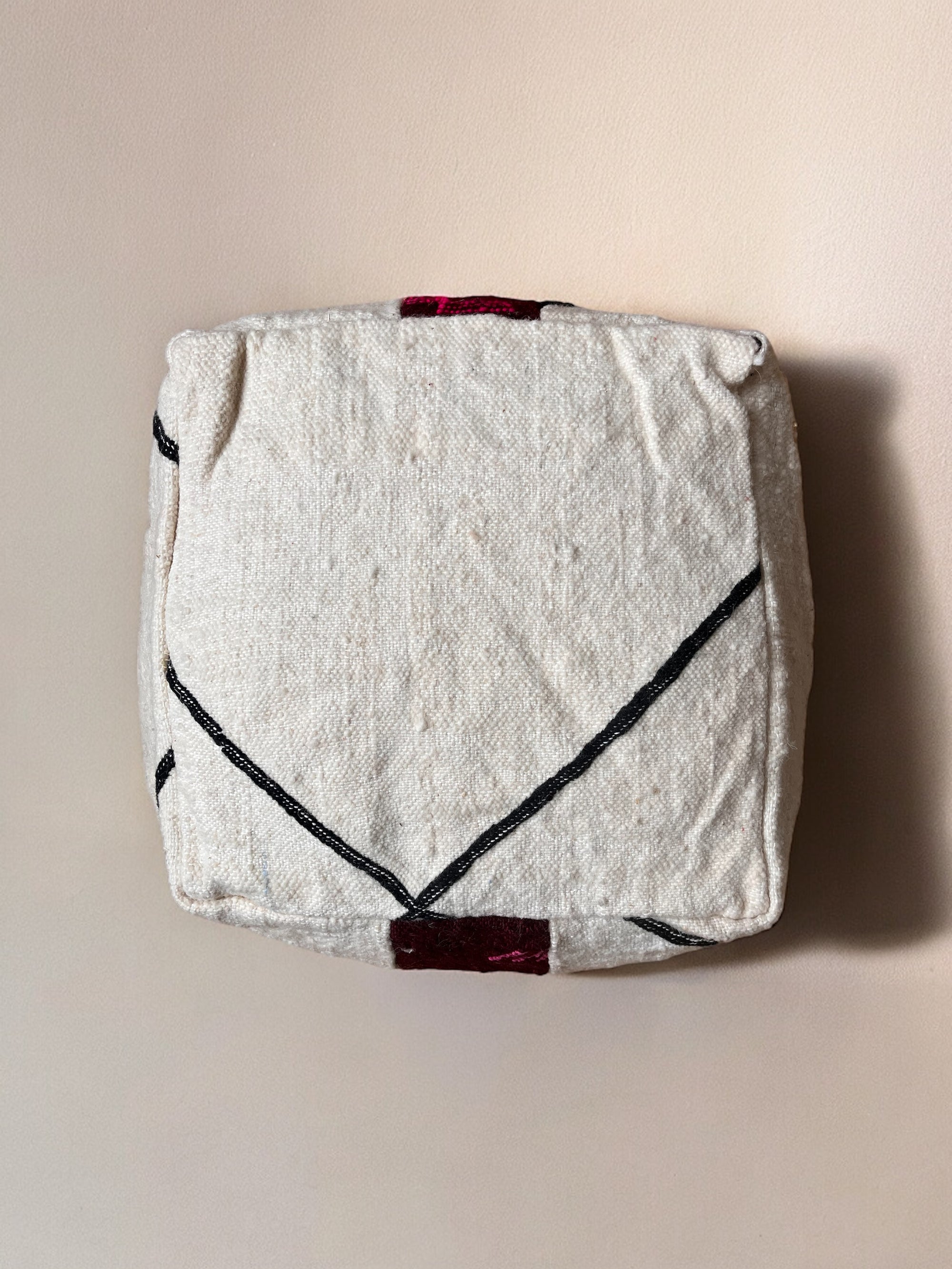Das Kleine Bodenkissen - Mera Mini von LEY! ist ein quadratisches, cremefarbenes Bodenkissen aus Schurwolle mit schwarzen diagonalen Linien und kleinen dunkelroten Akzenten, handgefertigt und auf einer hellbeigen Oberfläche abgebildet.