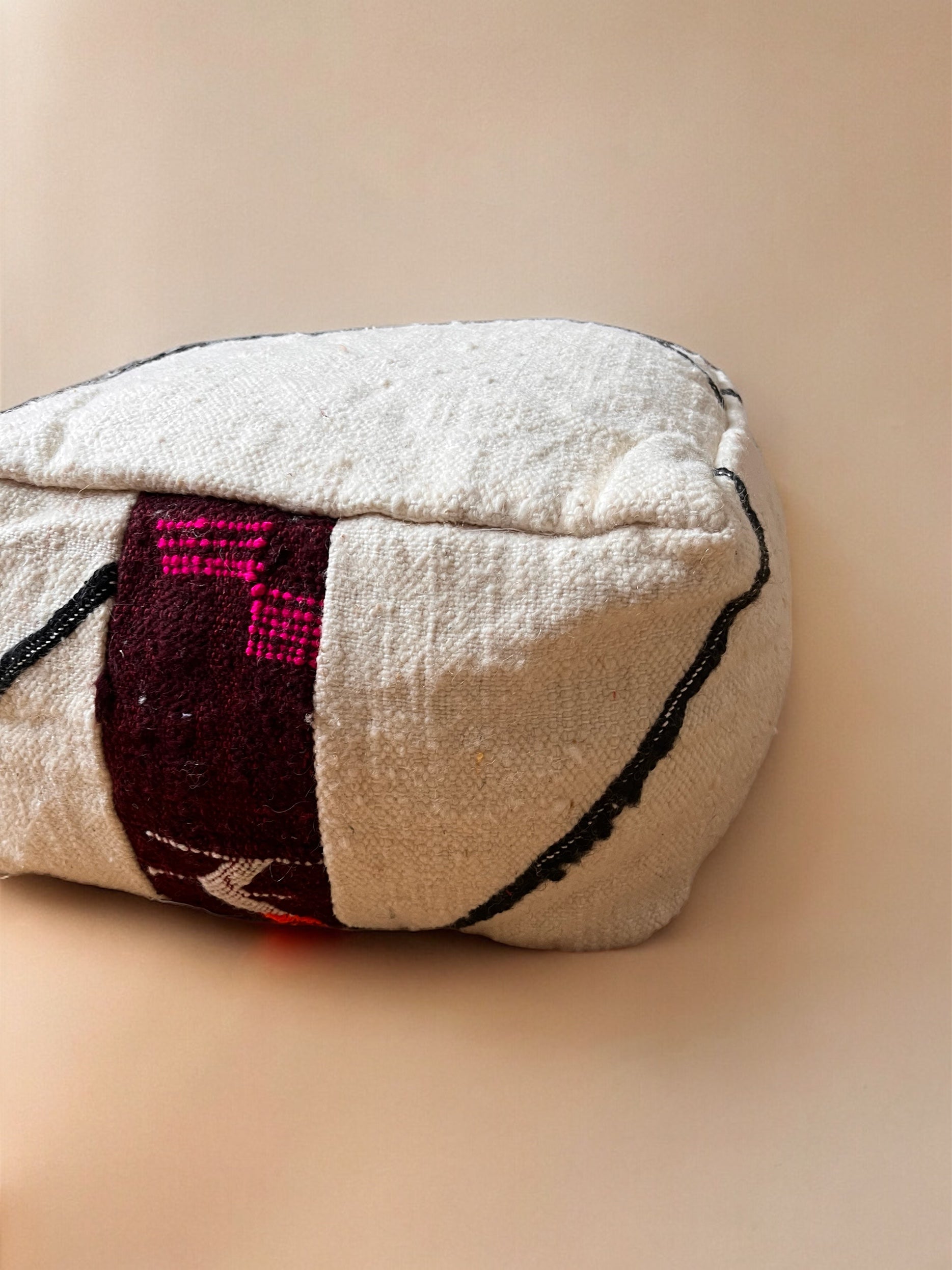 Das LEY! Kleines Bodenkissen - Mera Mini ist ein rechteckiges, strukturiertes weißes Kissen aus Schurwolle, das auf einer Seite ein kastanienbraunes und schwarzes geometrisches Muster auf einem neutralen beigen Hintergrund aufweist.