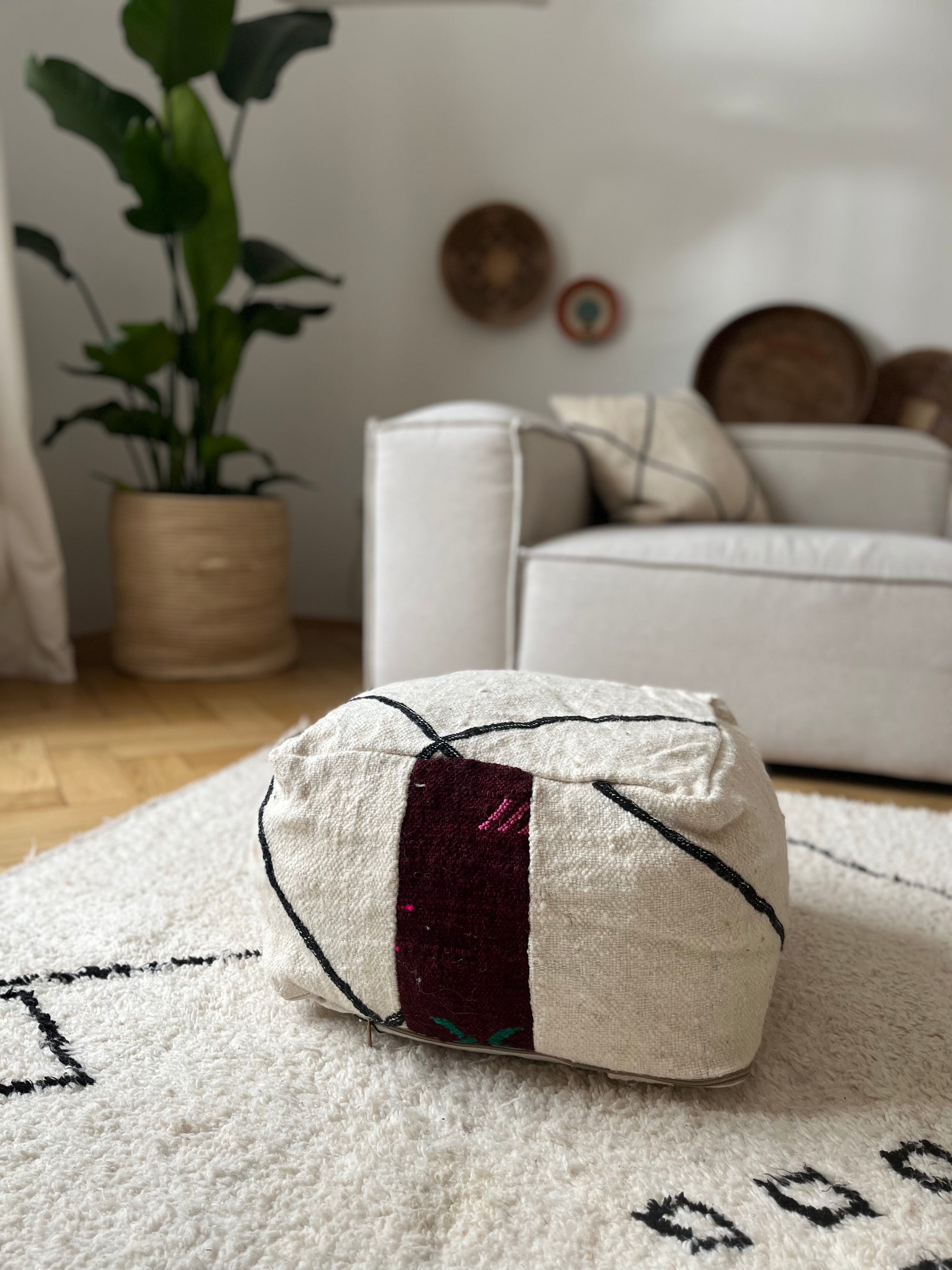 Das kleine Bodenkissen - Mera Mini von LEY! in Creme mit maronen- und schwarzen geometrischen Mustern liegt auf einem weißen Teppich in einem gemütlichen Wohnzimmer, dekoriert mit handgefertigter Wanddeko und einer großen Zimmerpflanze.