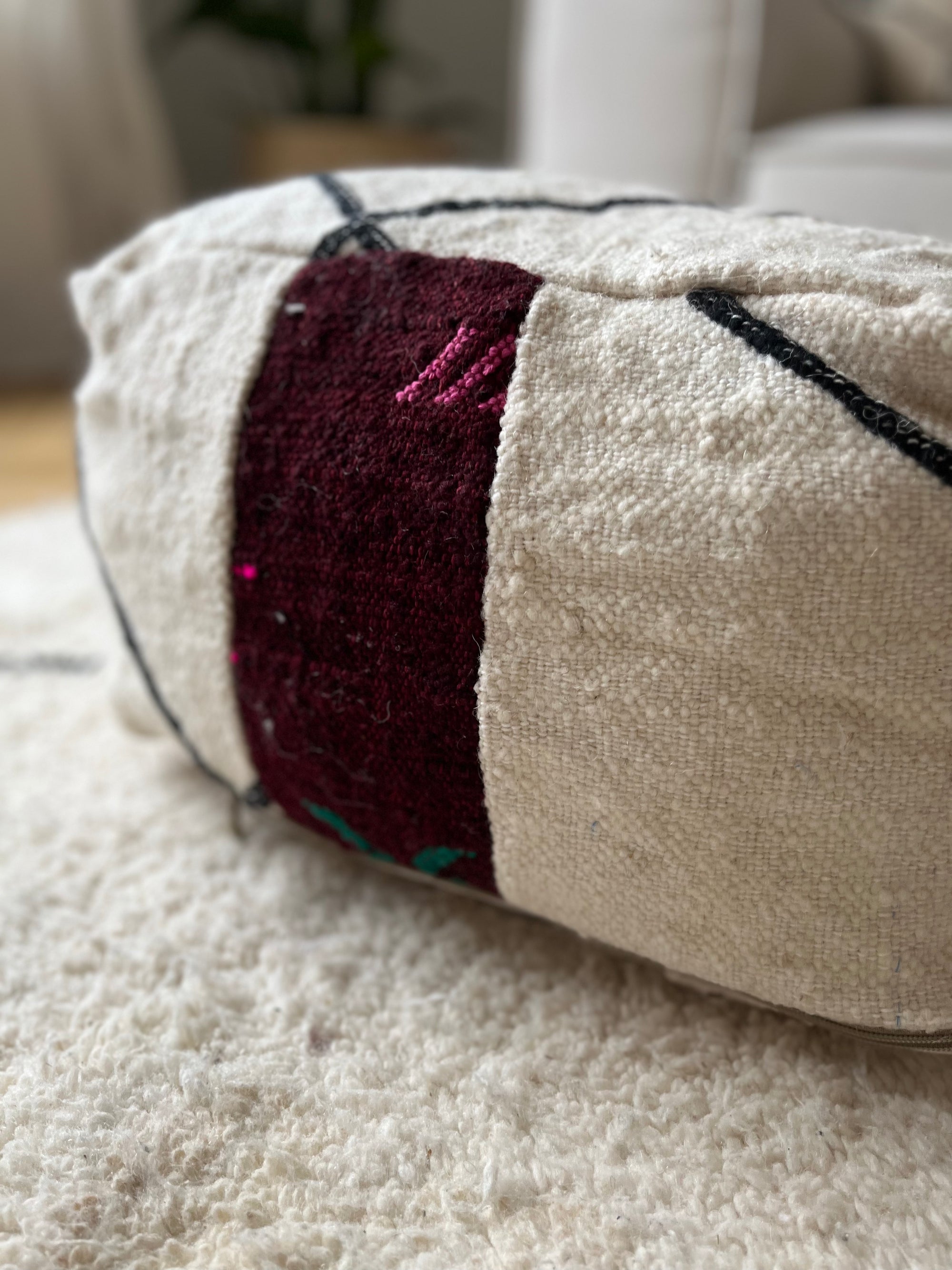 Eine Nahaufnahme des cremefarbenen Kleinen Bodenkissens - Mera Mini von LEY! mit einem dunkelroten rechteckigen Aufnäher und schwarzen Linien, das auf einem weichen weißen Teppich liegt. Der sanft verschwommene Hintergrund zeigt Andeutungen von Möbeln und einer Pflanze.