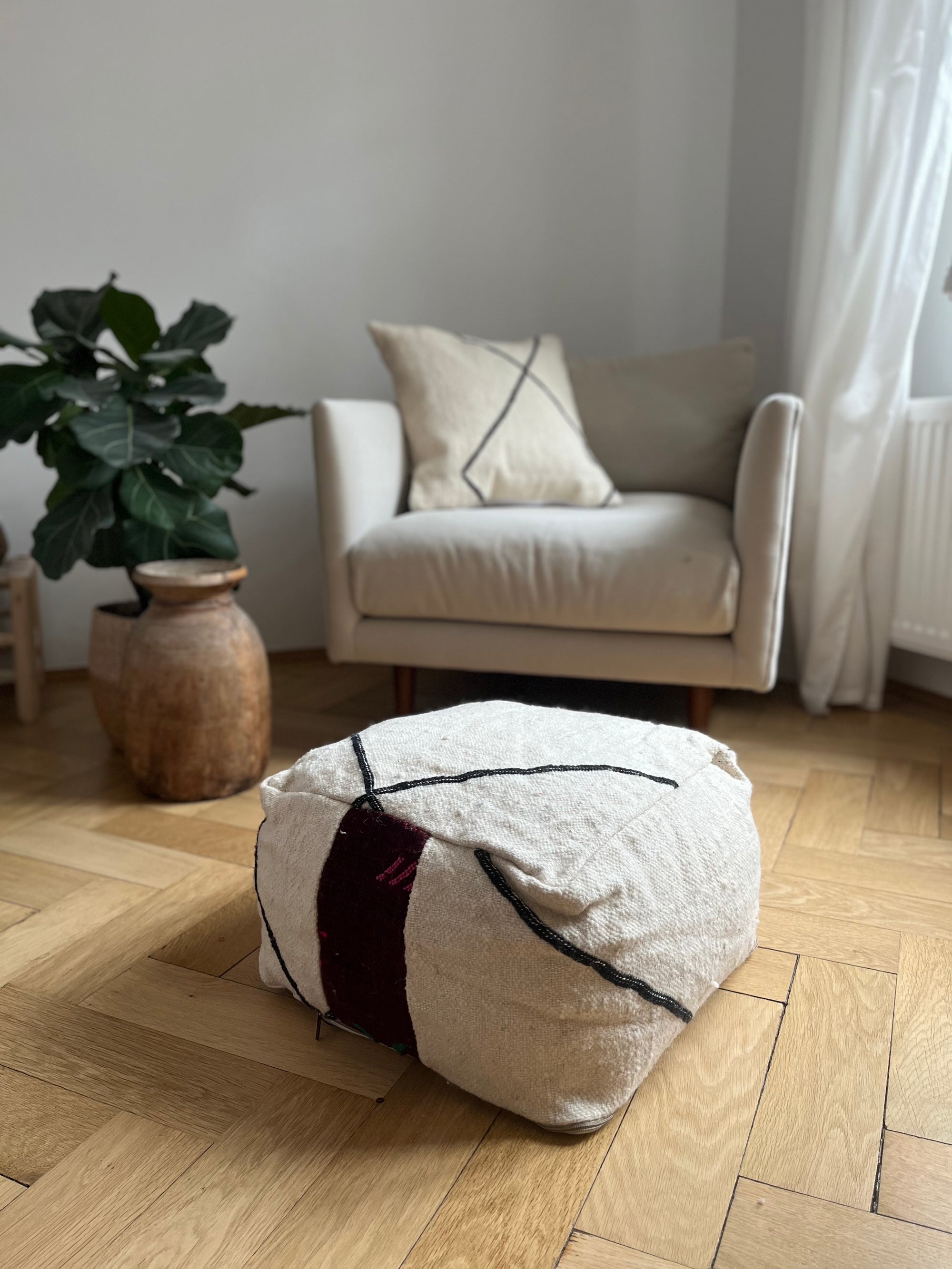 Ein gemütliches Wohnzimmer mit einem cremefarbenen Sessel, passendem Kissen, Topfpflanze, rustikaler Vase und dem LEY! Kleines Bodenkissen - Mera Mini aus reiner Schurwolle mit schwarzen und bordeauxfarbenen Streifen auf dem Holzboden.