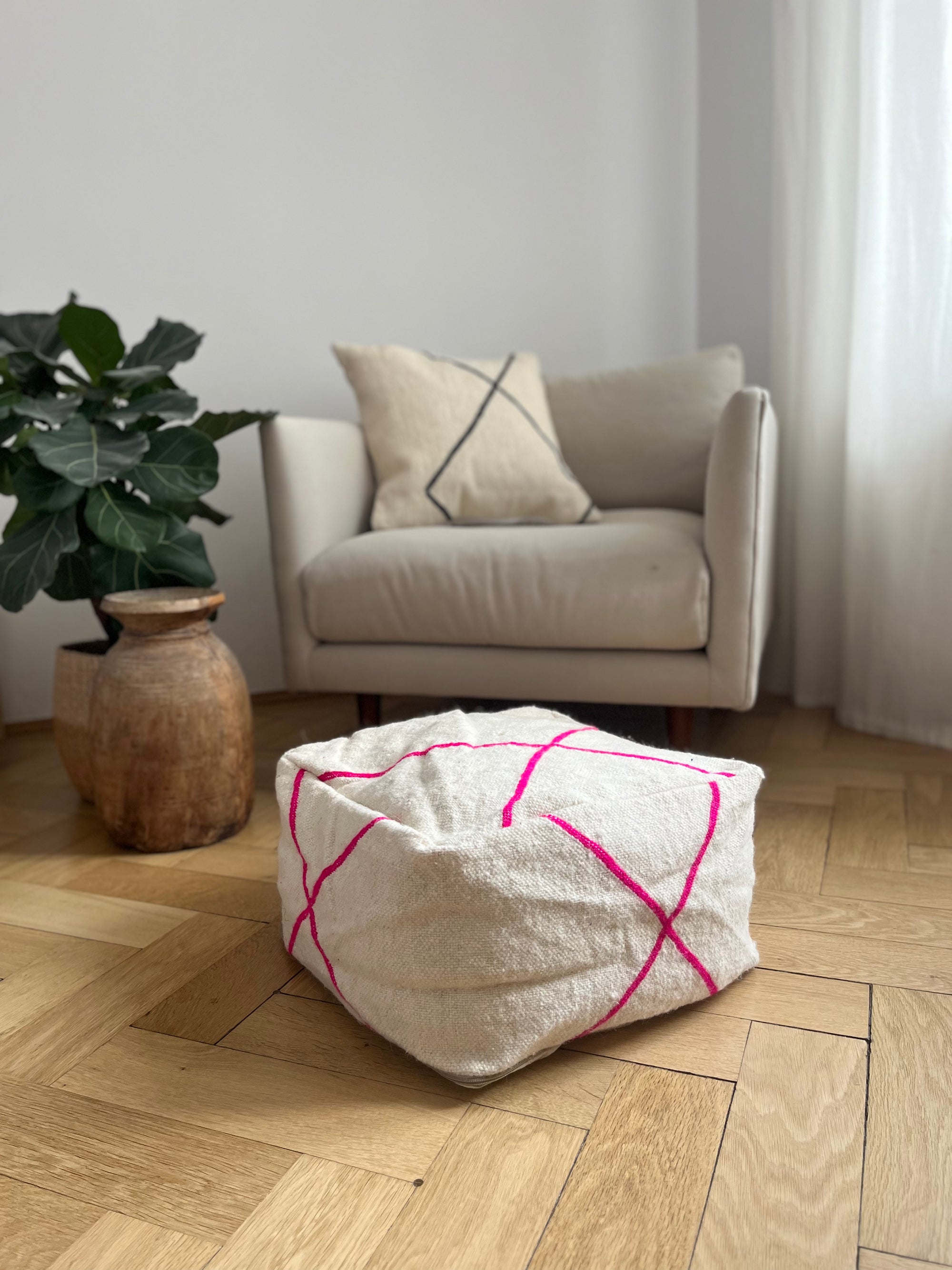 Ein cremefarbener Sessel mit beigem Kissen steht neben einer Blattpflanze in einem Keramiktopf. Im Vordergrund liegt das Kleines Bodenkissen - Mina Mini von LEY! mit leuchtend pinken Linien auf dem Holzboden.