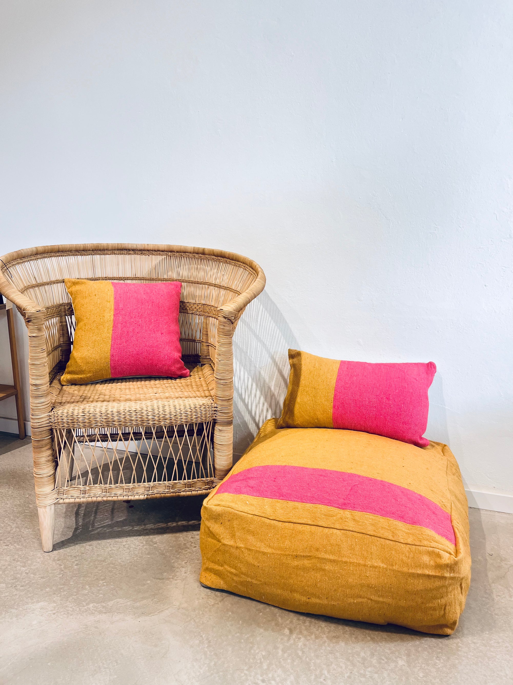 Ein Korbsessel und ein Pouf sitzen nebeneinander, beide mit LEY! Kissen - Alinya 40x40 in senfgelben und hellrosa Blöcken - die perfekte Frühlingsdekoration vor einer weißen Wand und einem polierten Betonboden.