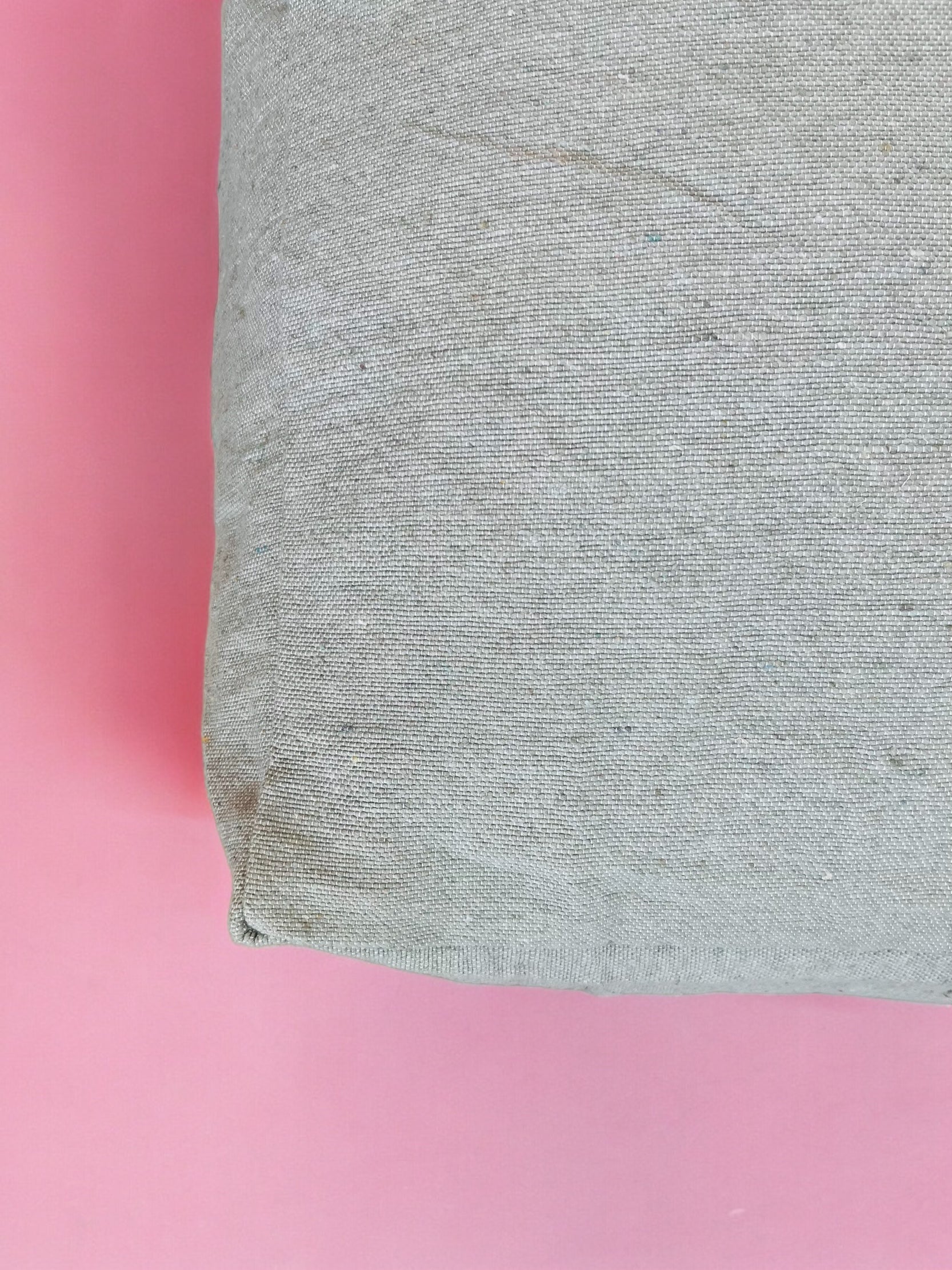 Eine Nahaufnahme des LEY! Bodenkissen - Milena, einem handgemachten, hellgrauen Baumwolle-Bodenkissen, platziert auf einer rosa Fläche mit dem Kissen auf der rechten Seite und dem rosa Hintergrund auf der linken Seite.