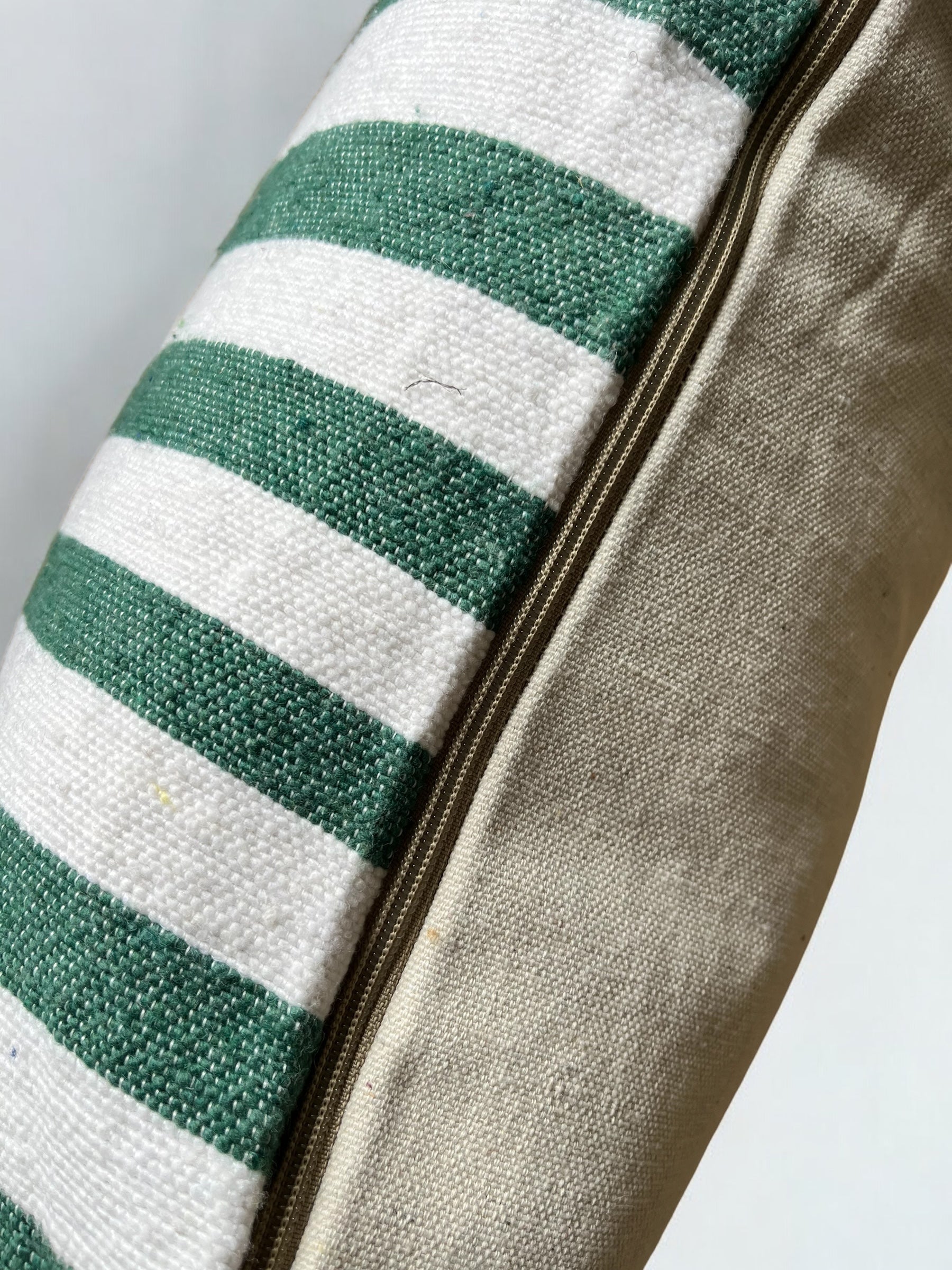 Nahaufnahme des LEY! Kissen - Tilosa 60x60, das auf der einen Seite grün-weiß gestreift und auf der anderen Seite einfarbig beige ist, getrennt durch einen sichtbaren Reißverschluss.