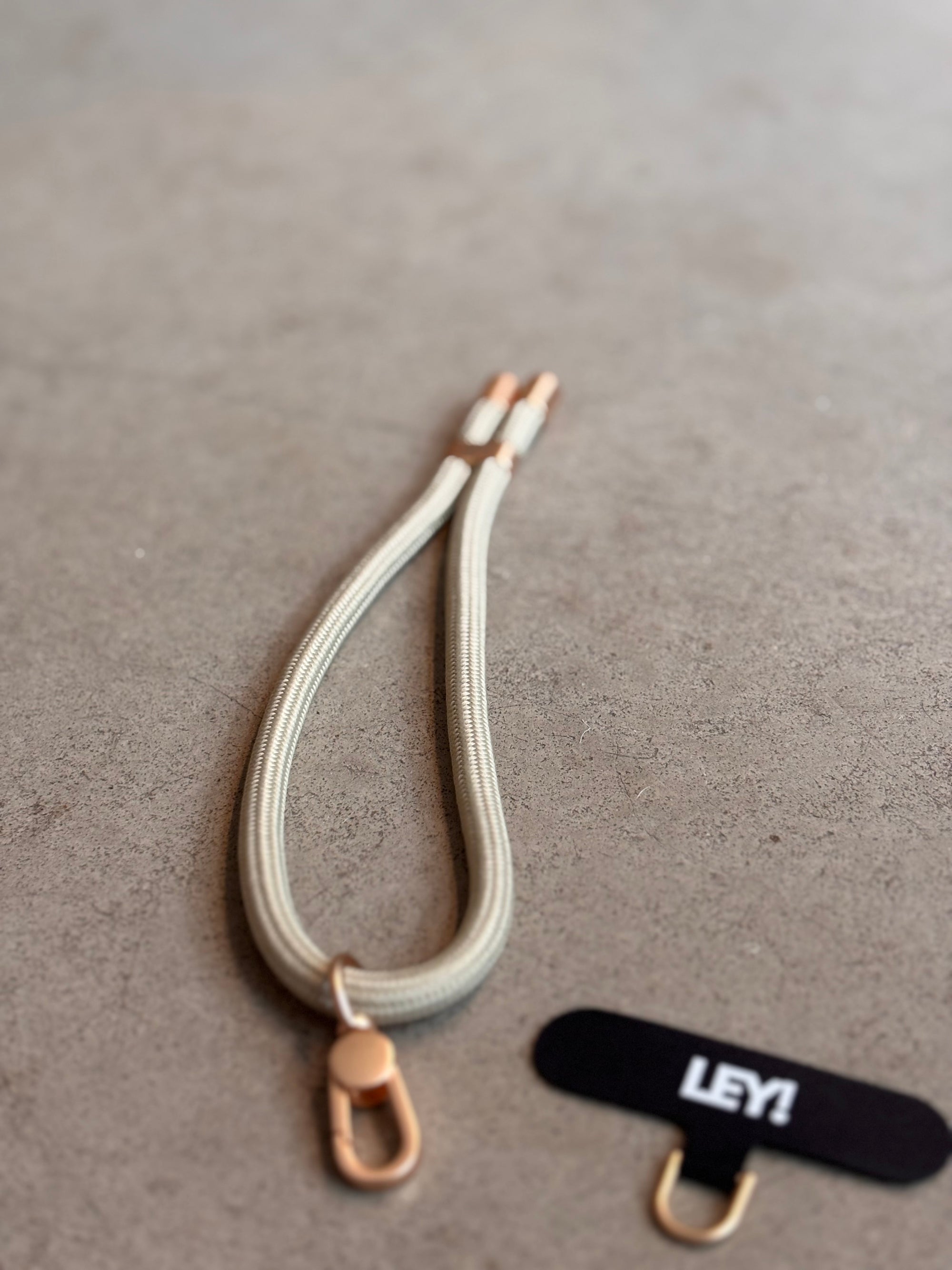 Ein beiger Schlüsselanhänger von LEY! mit goldfarbenen Clips und Karabiner liegt auf grauer Fläche neben einem schwarzen Label mit weißem "LEY!"-Schriftzug - praktisch, um deine Schlüssel immer griffbereit zu haben.