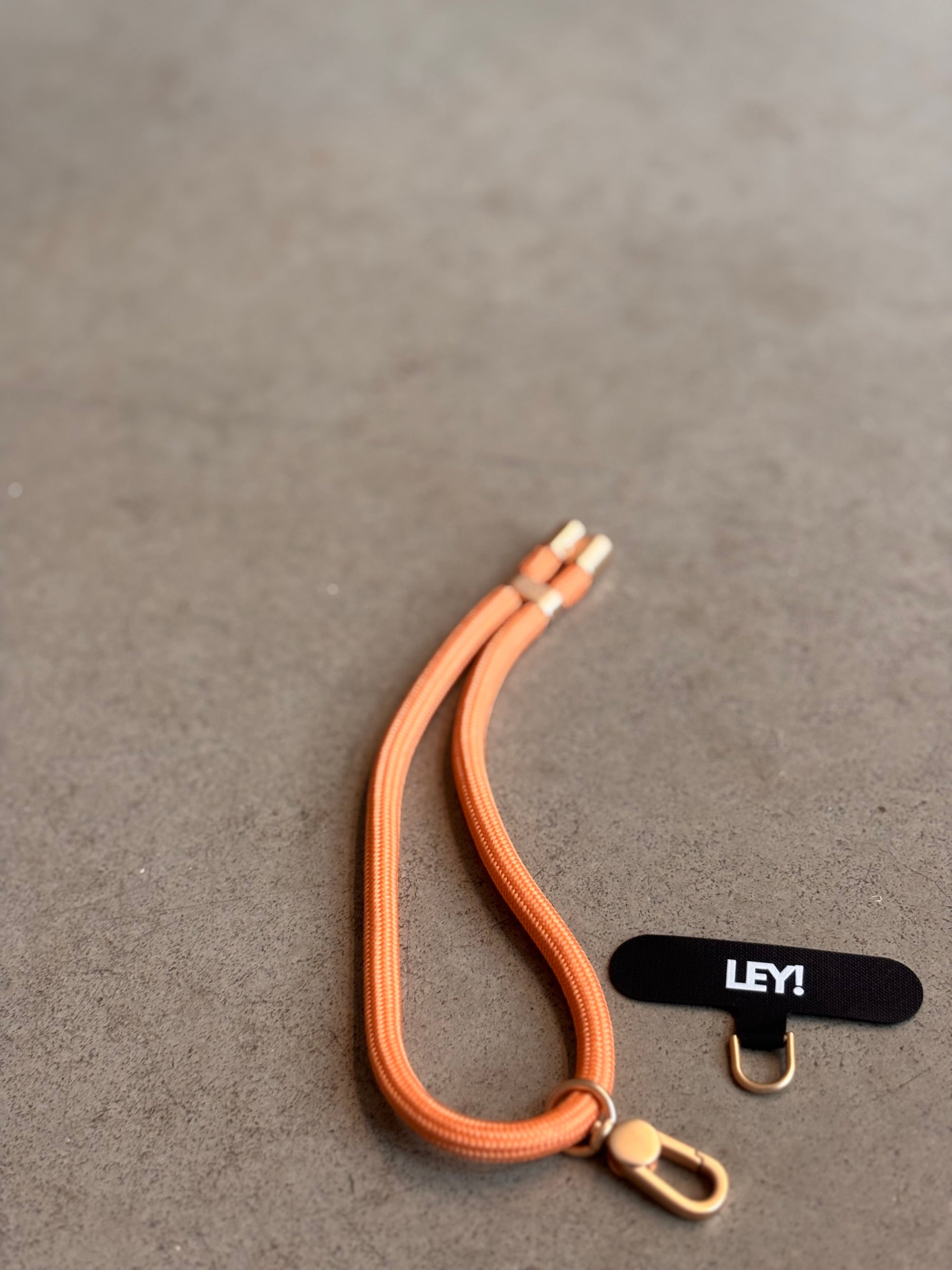 Ein leuchtend orangefarbener Schlüsselanhänger von LEY! mit goldenen Verschlüssen und einem goldenen D-Ring ruht auf einer grauen Fläche neben einem schwarzen Anhänger mit weißer Aufschrift "LEY!" und ist ein trendiges Accessoire für Ihre Schlüssel.